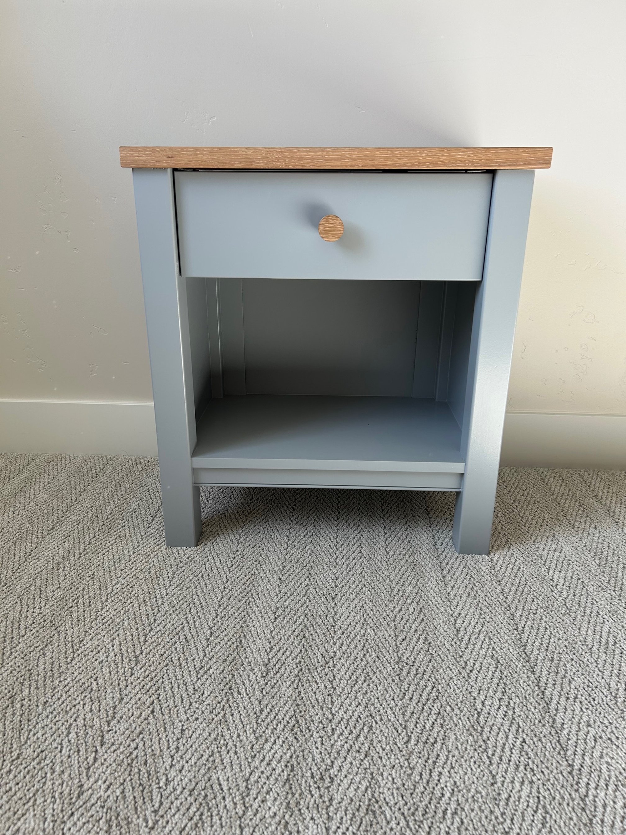 A blue nightstand with a single drawer and an open shelf, placed on a beige carpeted floor against a white wall.