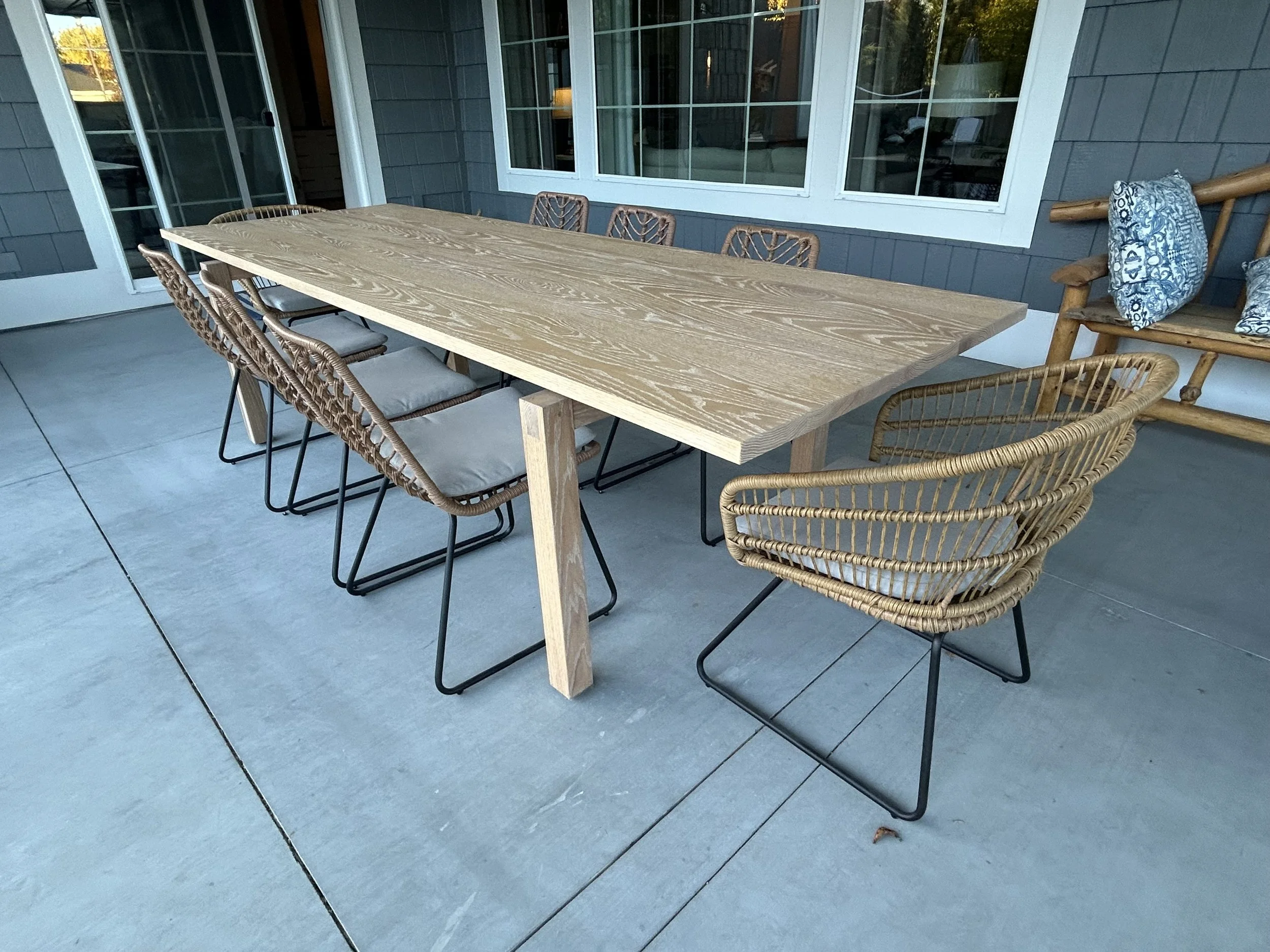 Outdoor patio table with eight woven chairs and a wooden bench with cushions, set on a concrete patio outside a house with gray siding and large windows.