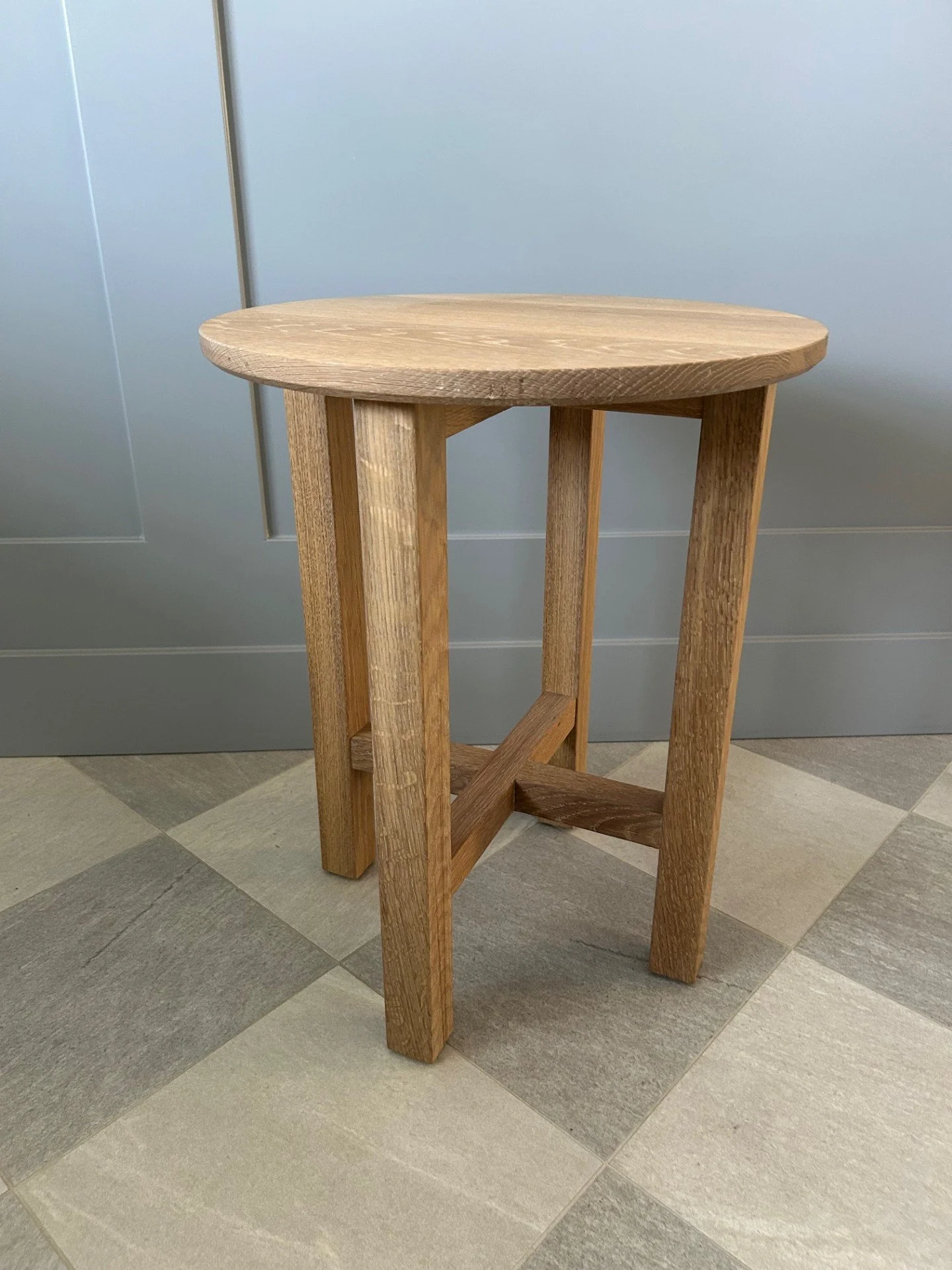 A wooden side table with a round top and straight legs, placed on a tiled floor against a light gray wall.