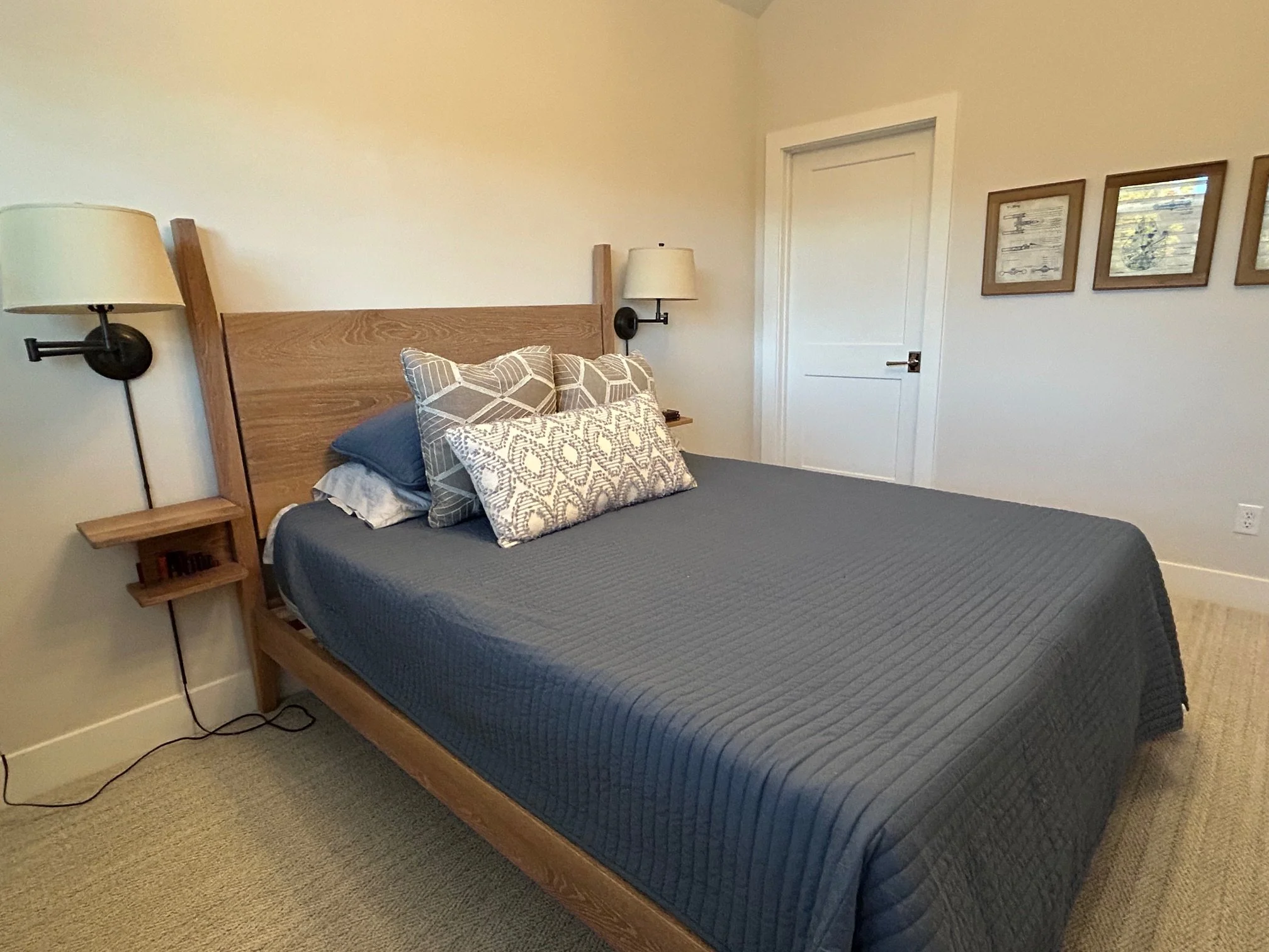 A neatly made bed with a wooden headboard, decorated with multiple patterned pillows and a dark blue quilt, in a minimalistic bedroom with wall-mounted lamps and framed artwork on the white walls.