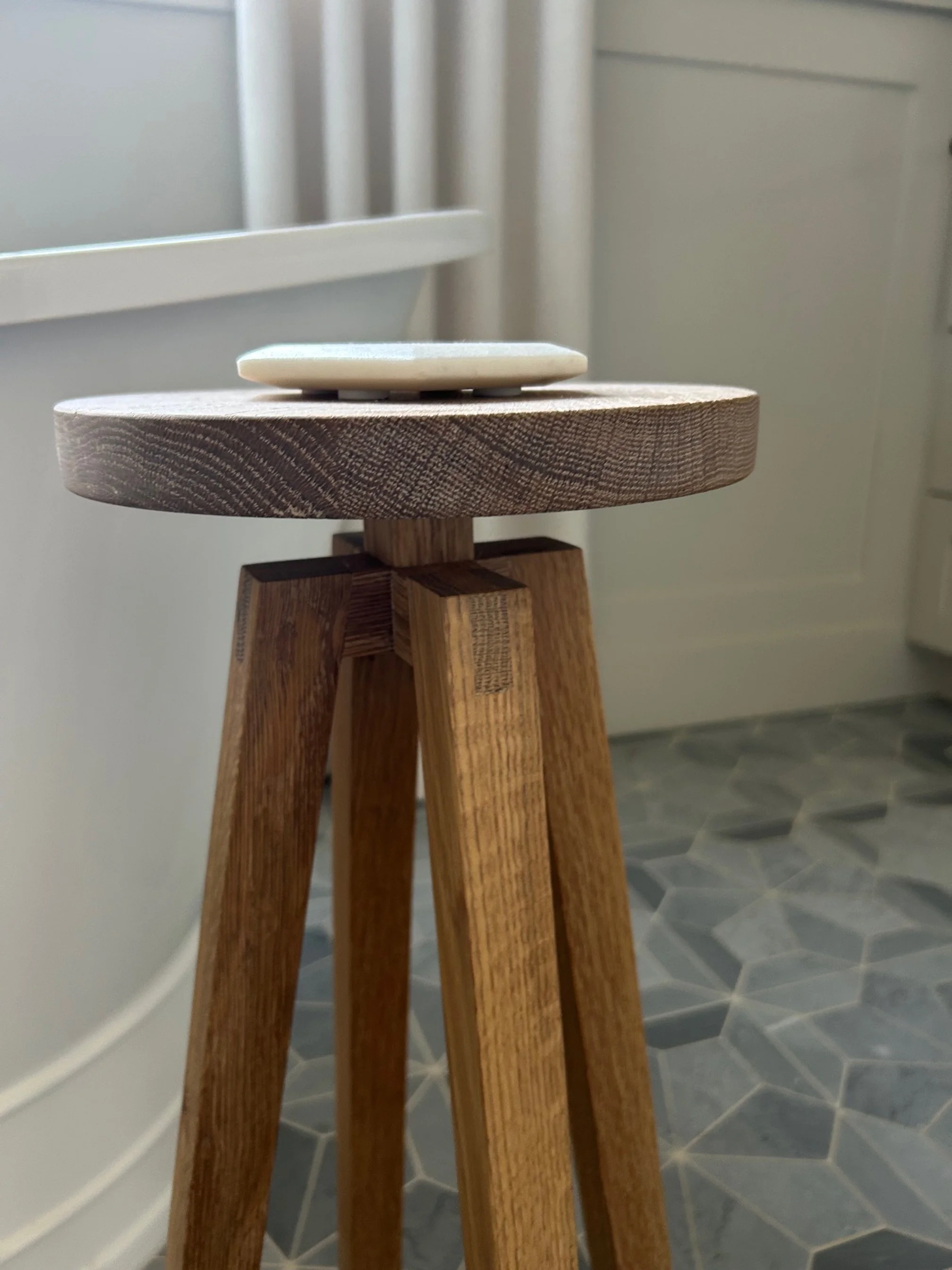 Close-up of a small wooden tripod with a ceramic dish on top, placed on a gray tiled floor near a white wall and radiator.