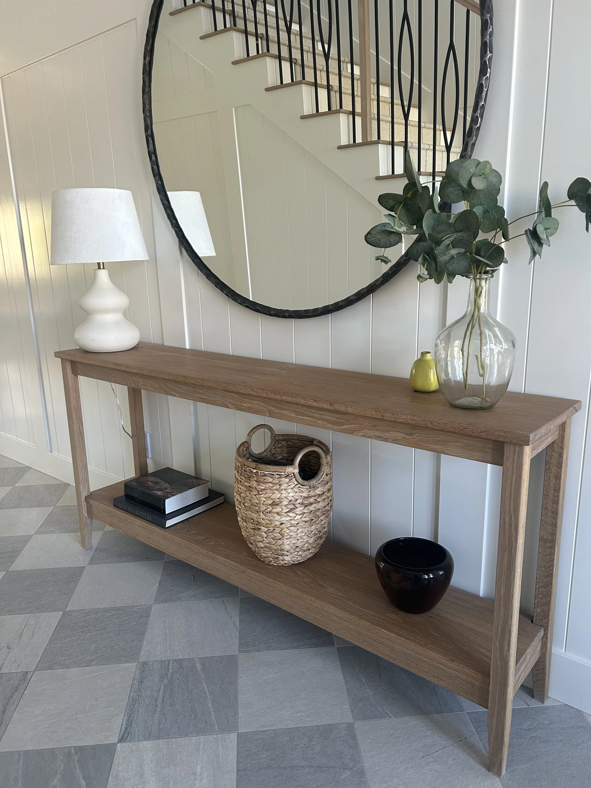 A wooden console table with a white table lamp, a large glass vase with green leaves, a small yellow vase, a woven basket, a black ceramic bowl, and two stacked books. A large round mirror is mounted on the wall above the table, reflecting stairs and