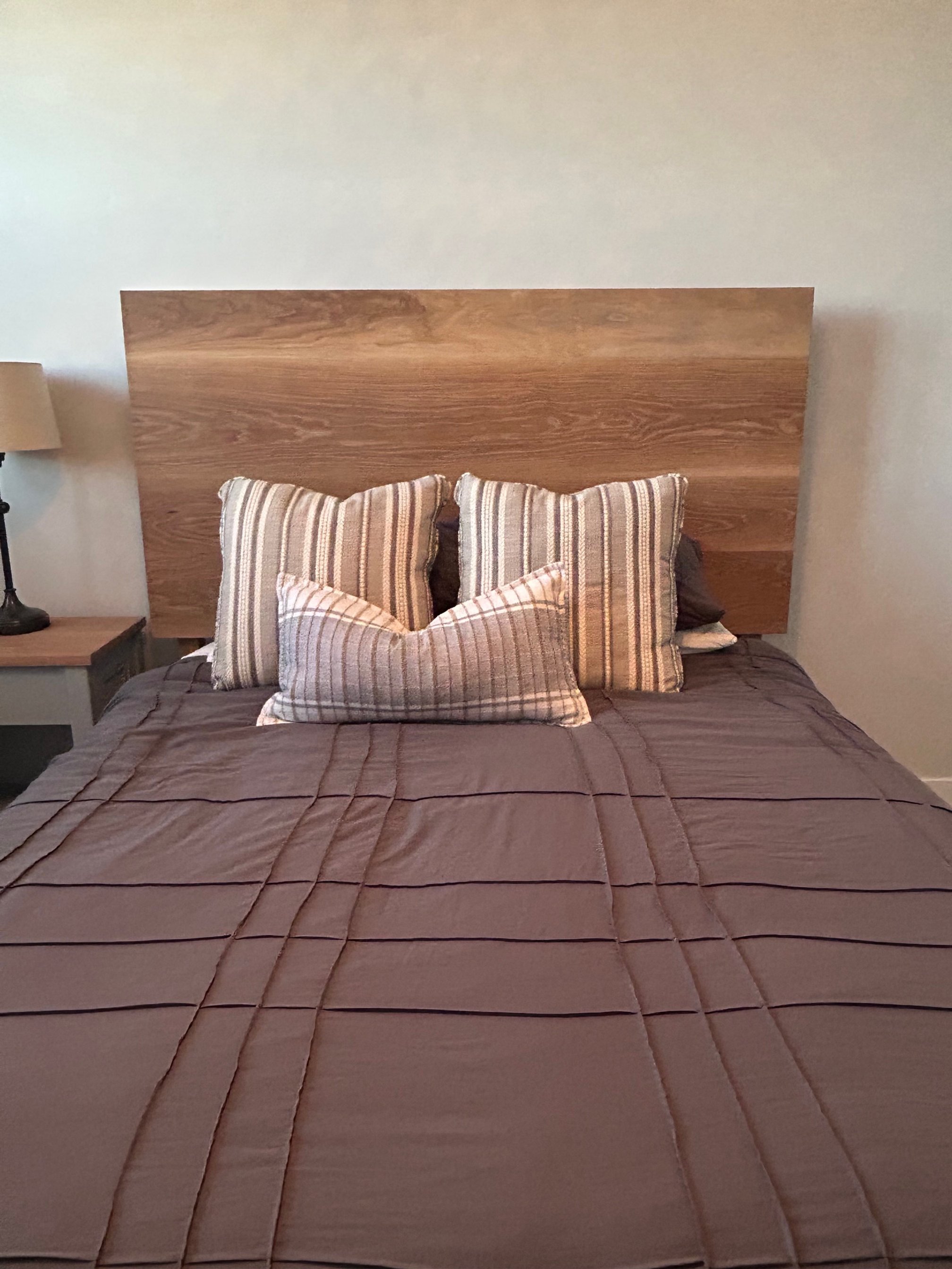 A neatly made bed with a wooden headboard, brown bedspread, and four striped pillows in beige, brown, and white.