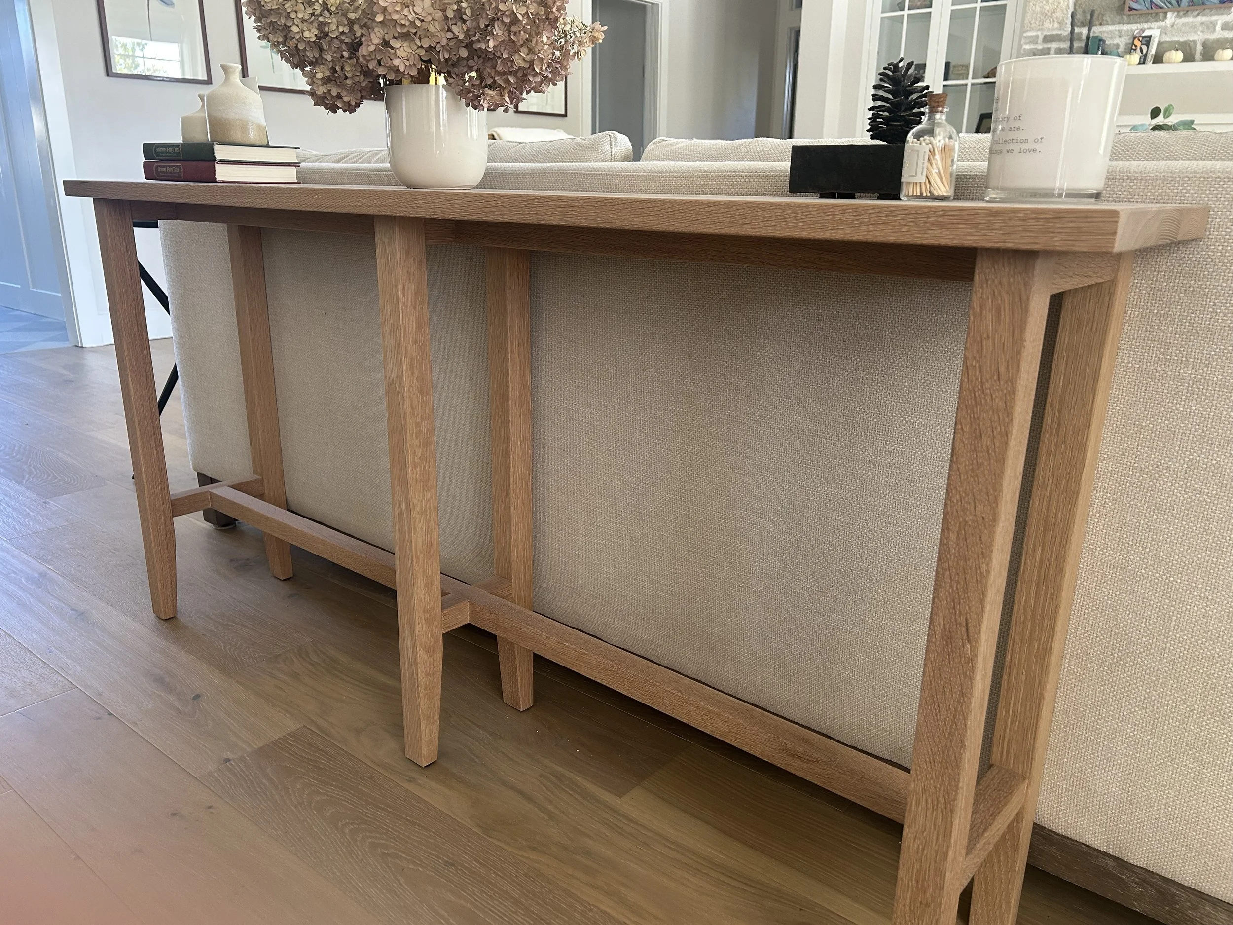 Wooden table upside down with decorative items on top, including a vase with dried flowers, a pinecone, a small jar, and candles, in a living room.
