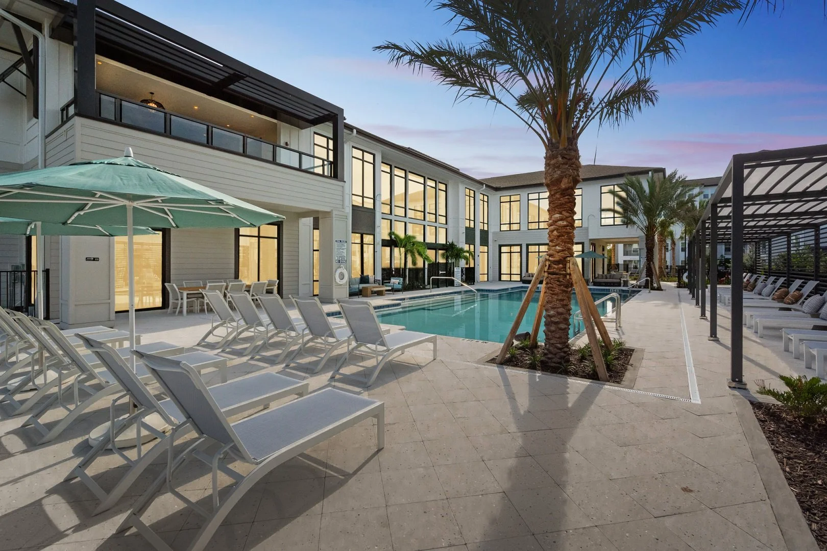 Outdoor pool area at a modern residential complex or hotel with lounge chairs, umbrellas, palm trees, and a building with large windows at dusk.