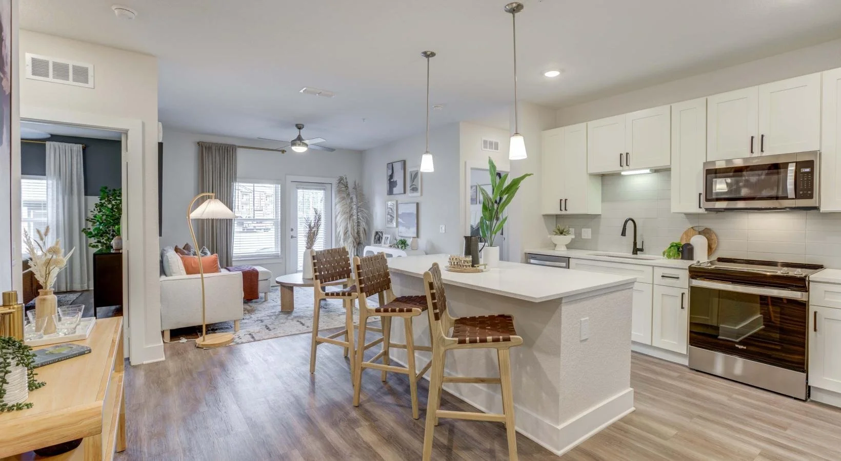Open-concept living room and kitchen with white cabinetry, stainless steel appliances, a large kitchen island with barstools, and cozy living area with sofa, curtains, and artwork.