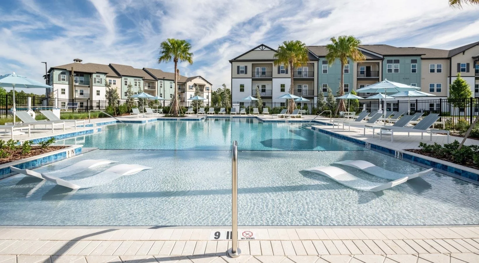 Outdoor swimming pool with lounge chairs, umbrellas, and palm trees surrounding a residential apartment complex on a sunny day.