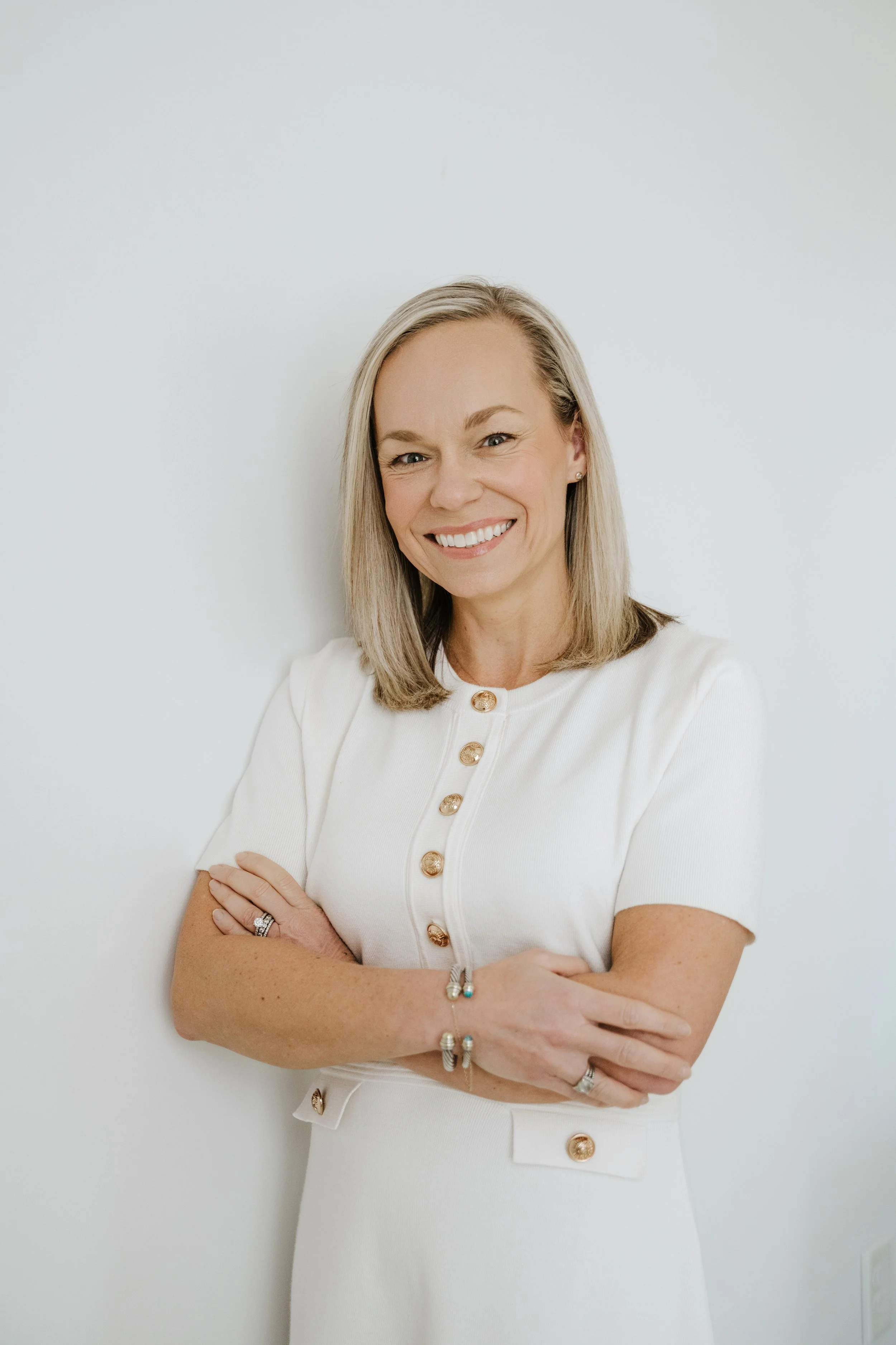 Portrait of a smiling woman with blonde hair, wearing a white dress with gold buttons, standing against a plain white background.