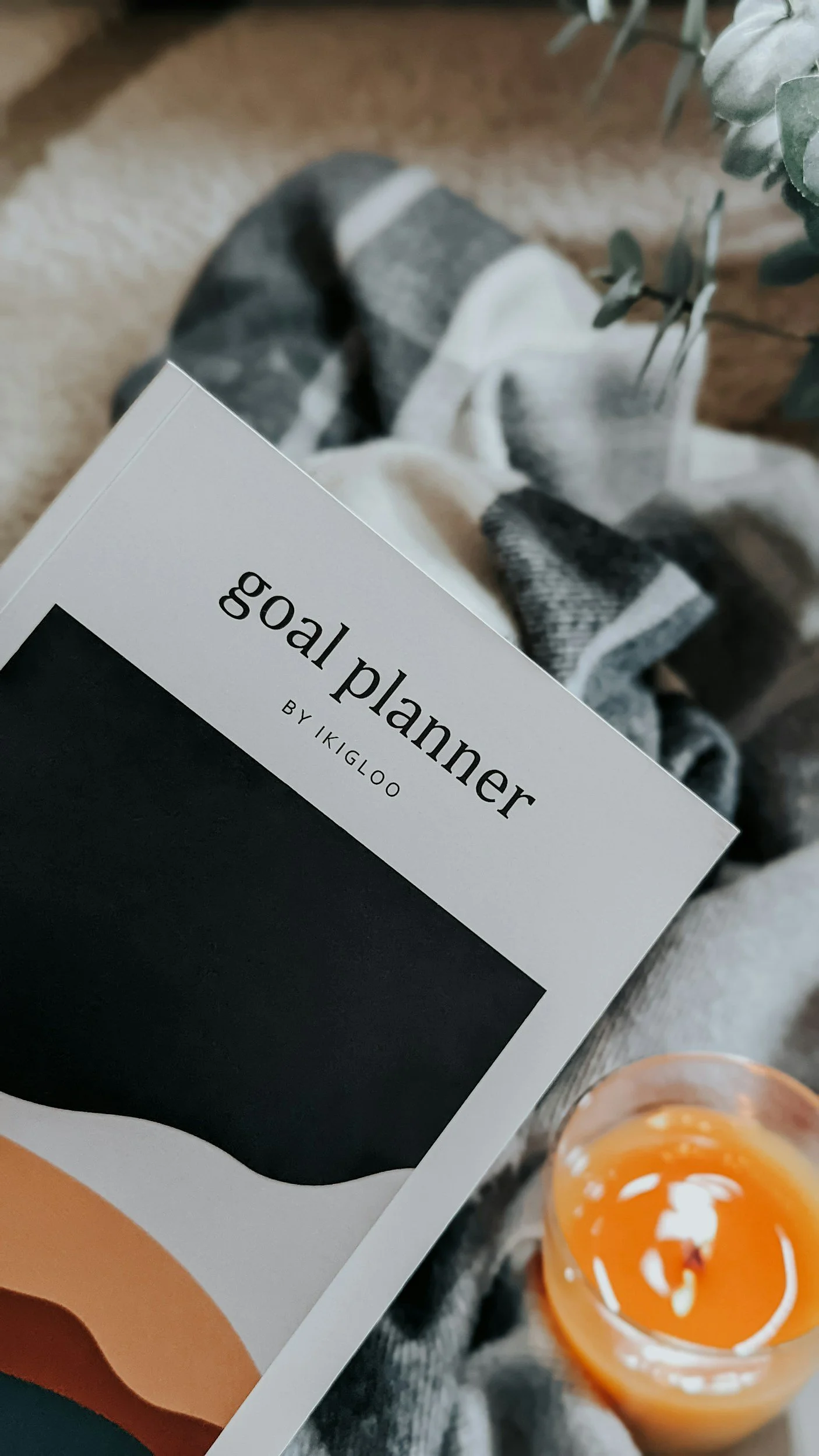 A person holding a book titled "goal planner" by Ikigloo, with a cup of orange-colored beverage nearby, sitting on a patterned blanket or carpet with some green plant leaves visible in the background.