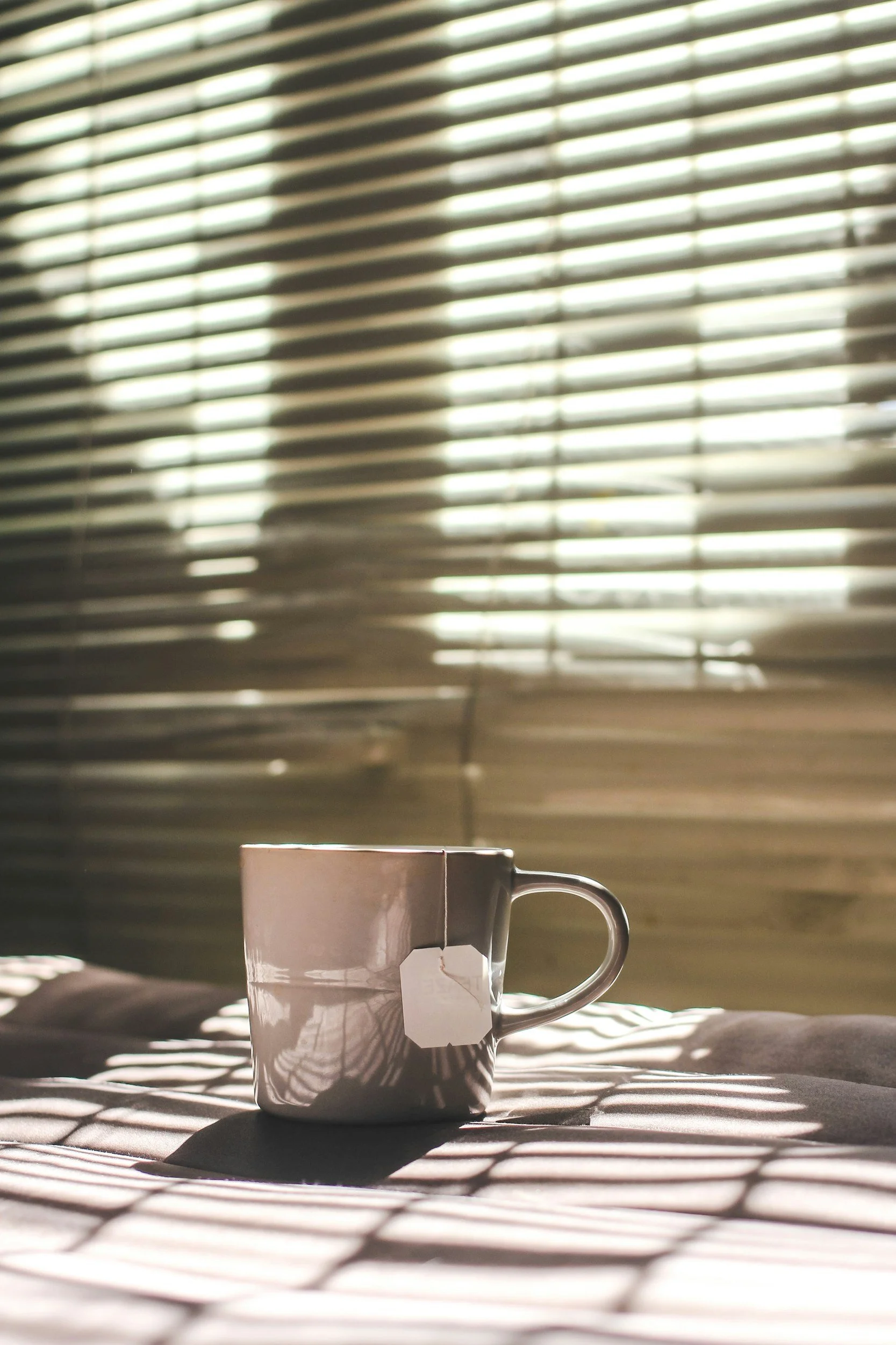 A white mug with a tea bag tag sitting on a bed, with sunlight streaming through window blinds, casting shadows on the bed and mug.