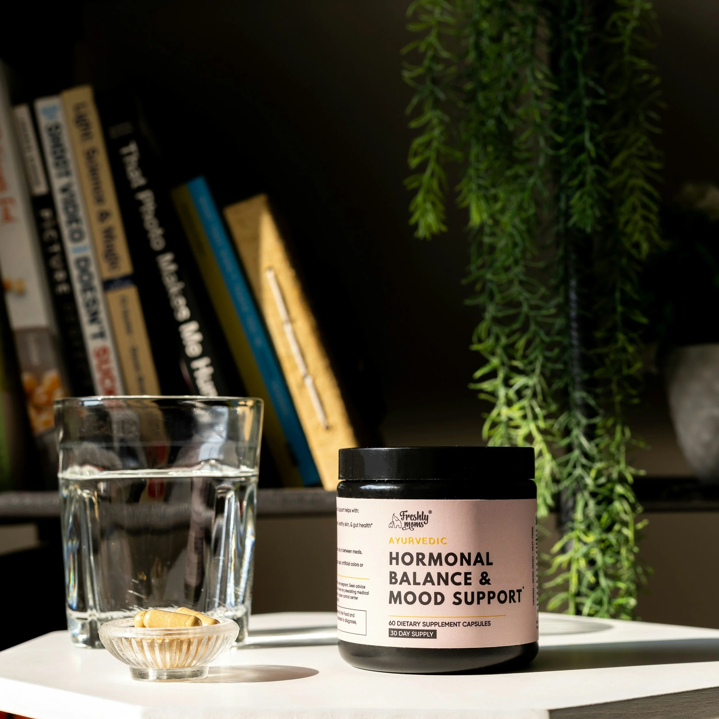 A jar of Ayurvedic dietary supplement capsules labeled 'Hormonal Balance & Mood Support' next to a glass of water and a dish with additional capsules, set on a white surface with a bookshelf and a green plant in the background.