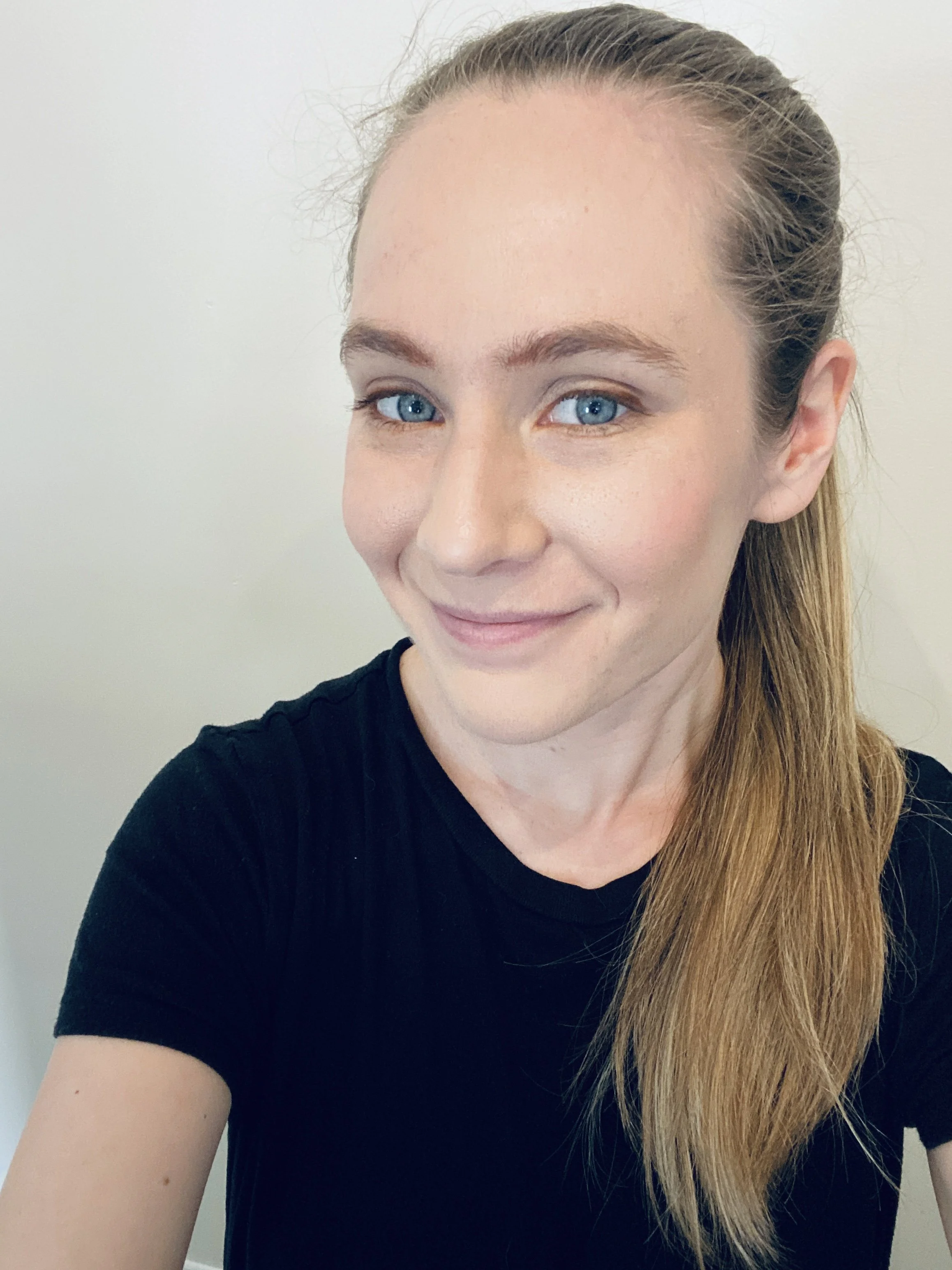 A woman with blonde hair tied back, wearing a black shirt, smiling, standing against a plain light-colored background.