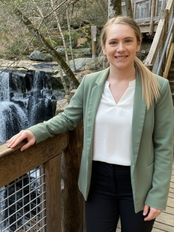 A young woman with long blonde hair smiling, standing next to a railing near a small waterfall in a wooded area.