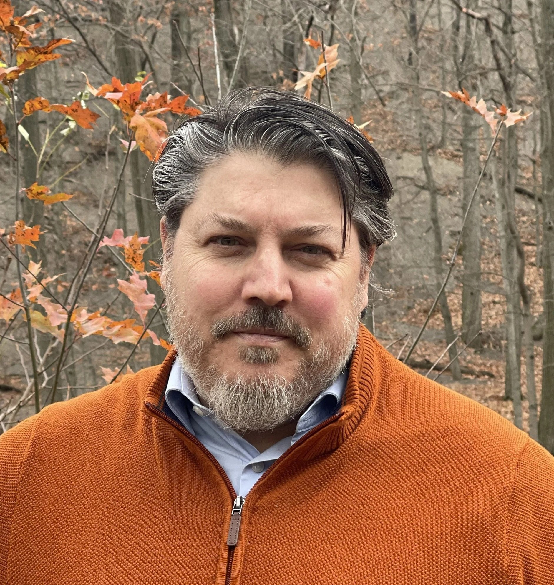 A man with gray hair and a beard wearing an orange jacket standing outdoors with leafless trees and some orange and pink fall leaves in the background.
