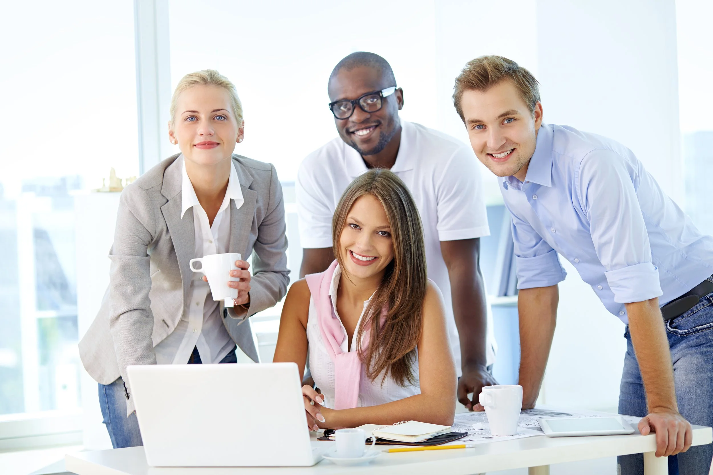 Group of five diverse young adults, three men and two women, gathered around a desk in an office, smiling at the camera with a laptop, notebooks, coffee cups, and a tablet in front of them.