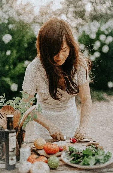 Woman cutting vegetables at outdoor kitchen table with fresh produce and salad.