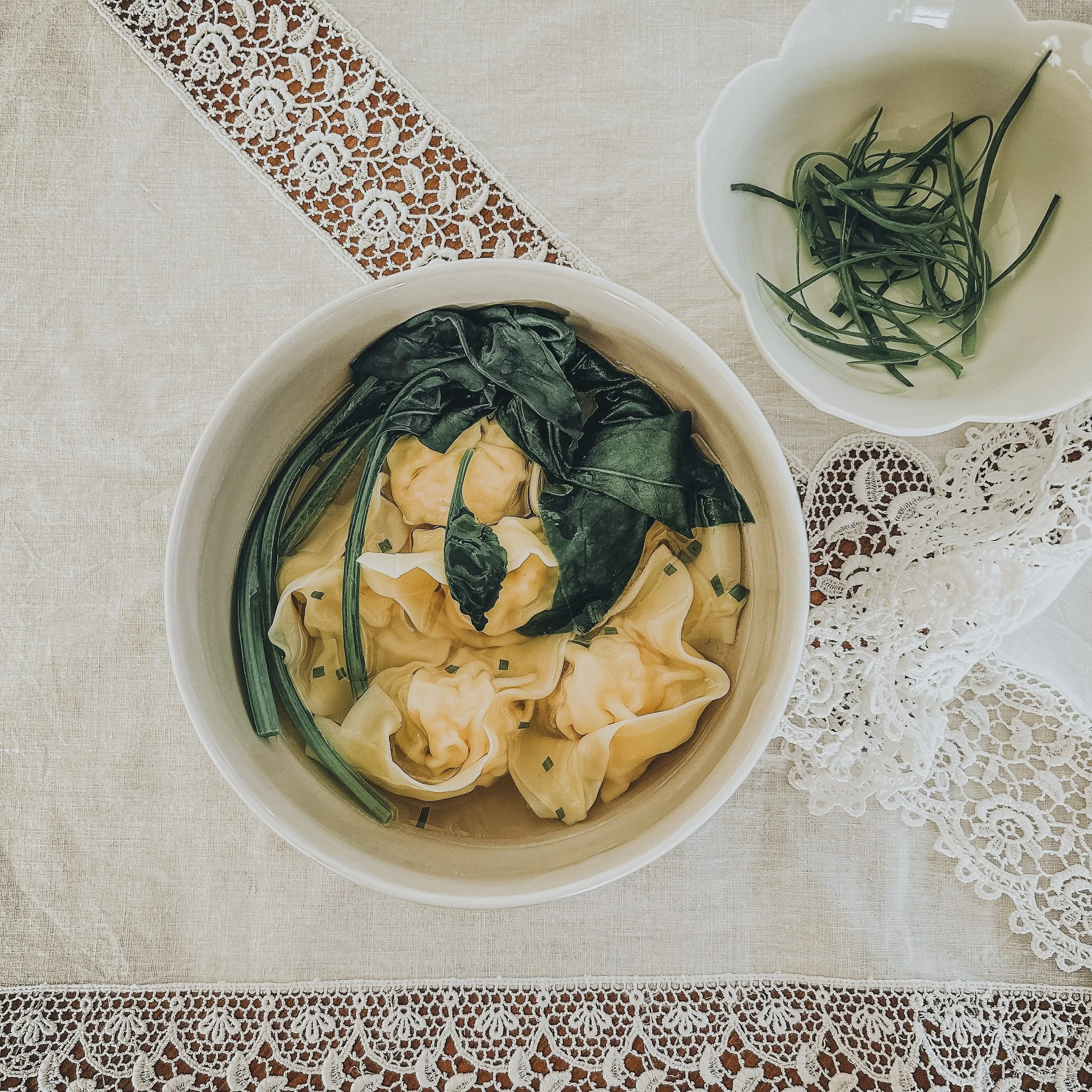A bowl of wonton soup with green leafy vegetables and chopped chives, accompanied by a smaller bowl of fresh chives on a lace tablecloth.