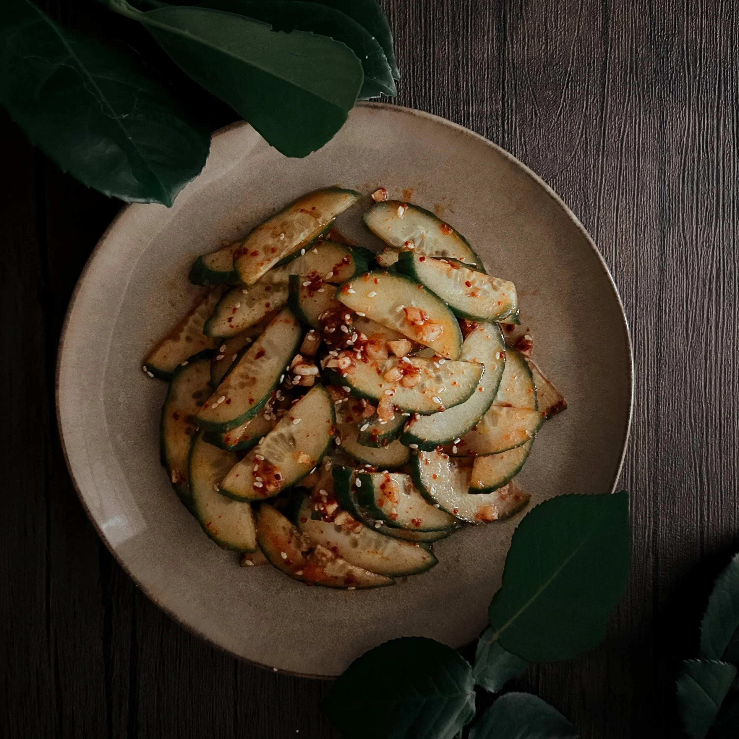 A plate of sliced cucumbers garnished with chopped nuts and red pepper flakes, on a beige dish surrounded by green leaves.