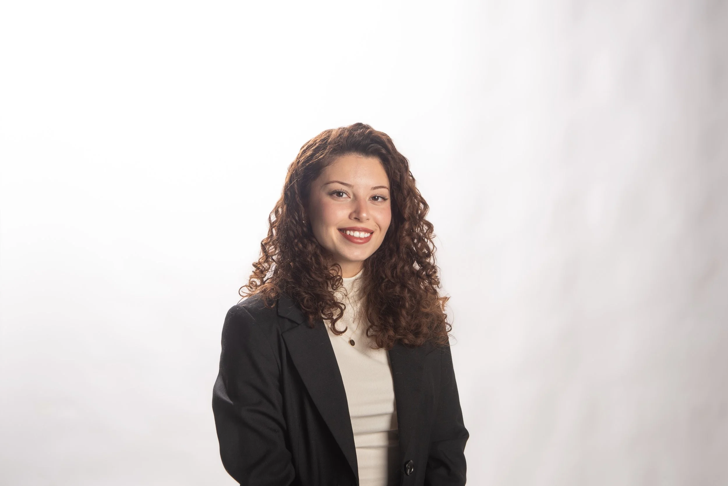 Portrait of a young woman with curly brown hair wearing a black blazer and cream-colored top, smiling against a plain white background.