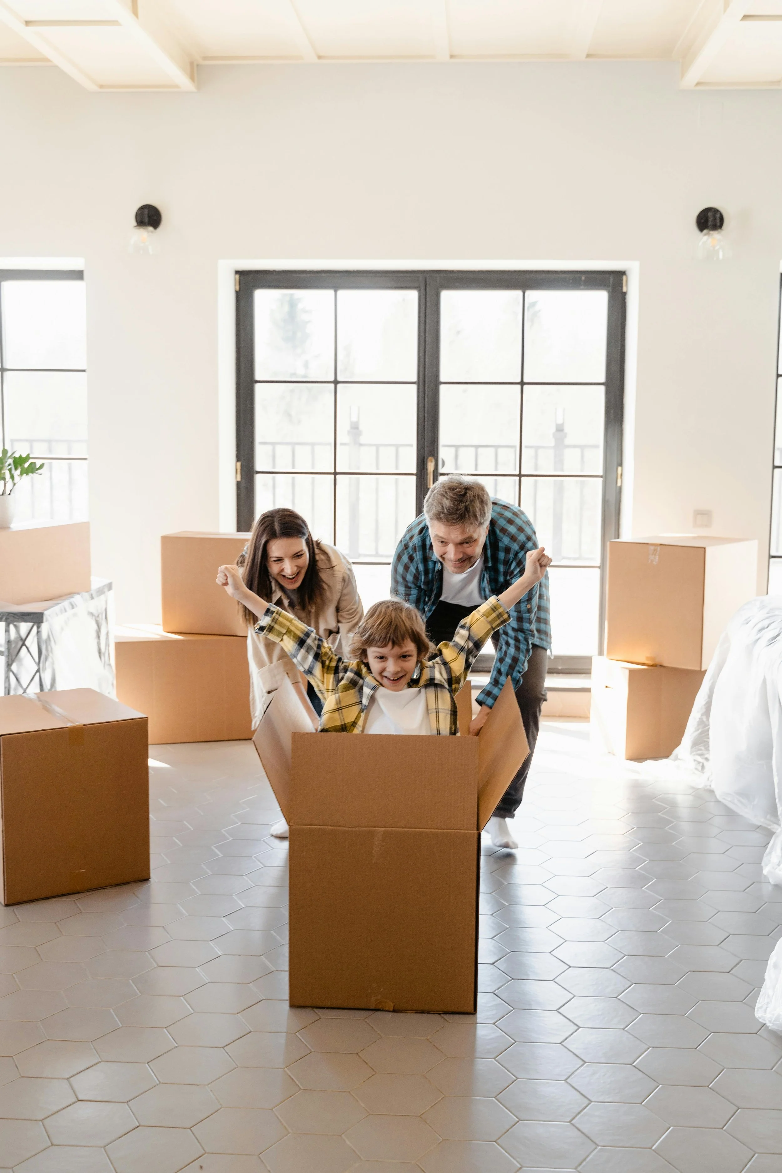 A family celebrating in a living room with moving boxes, one child excitedly sitting in an open cardboard box with arms raised, parents smiling and cheering.
