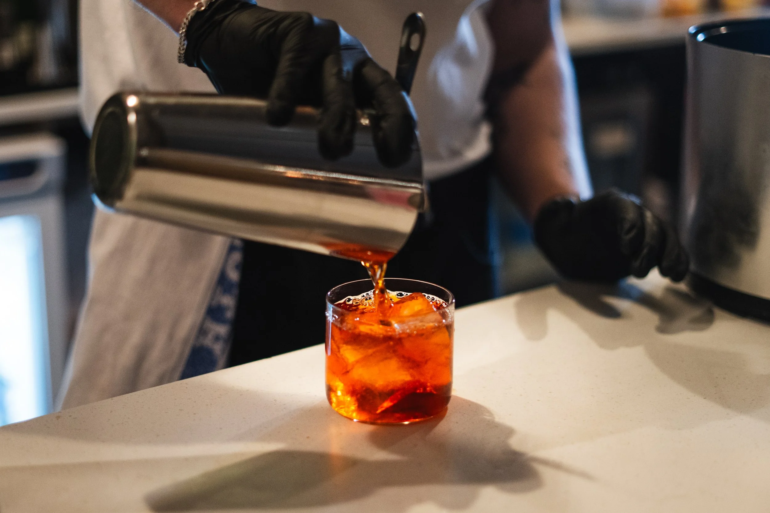 A person wearing black gloves pours a dark liquid from a shaker into a glass with ice, creating a cocktail.