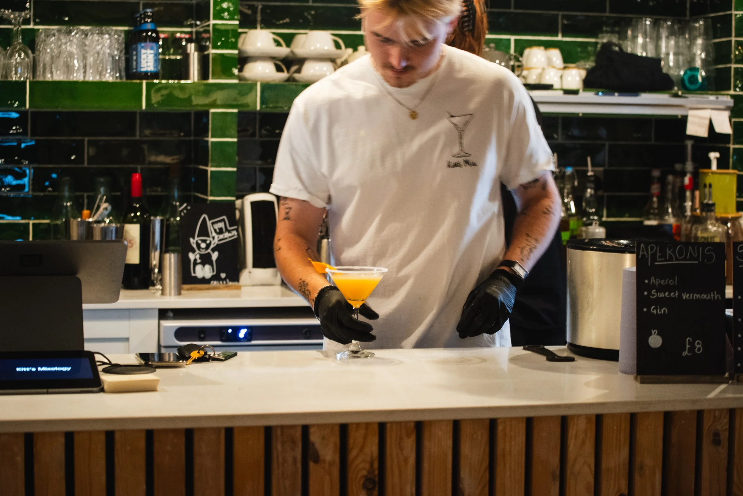 A bartender preparing a yellow cocktail in a martini glass behind a bar counter with various bottles and glasses, green tiled wall, and a blackboard menu.
