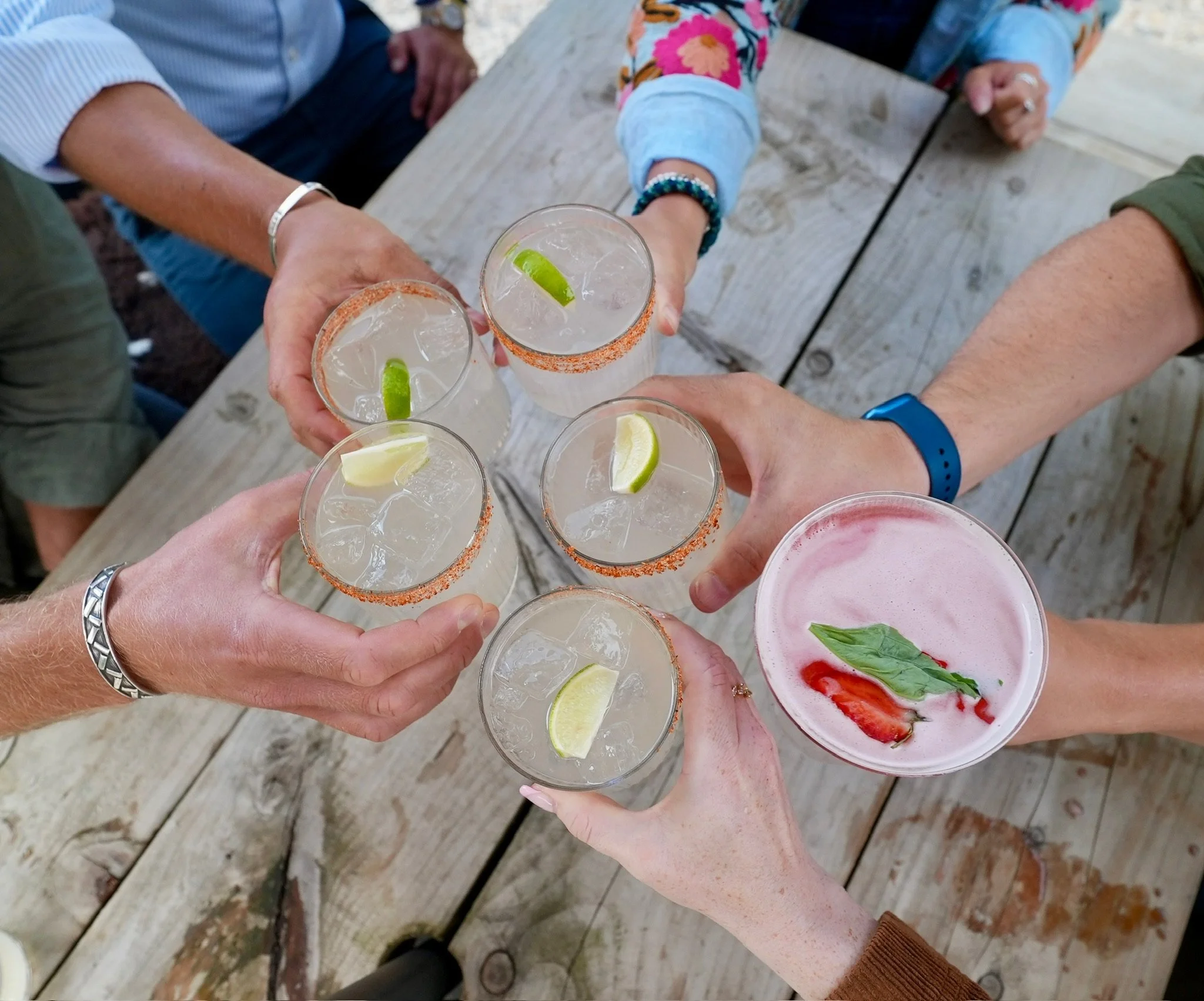 People holding up glasses of cocktails with lime and lemon wedges, toasting at a wooden table.