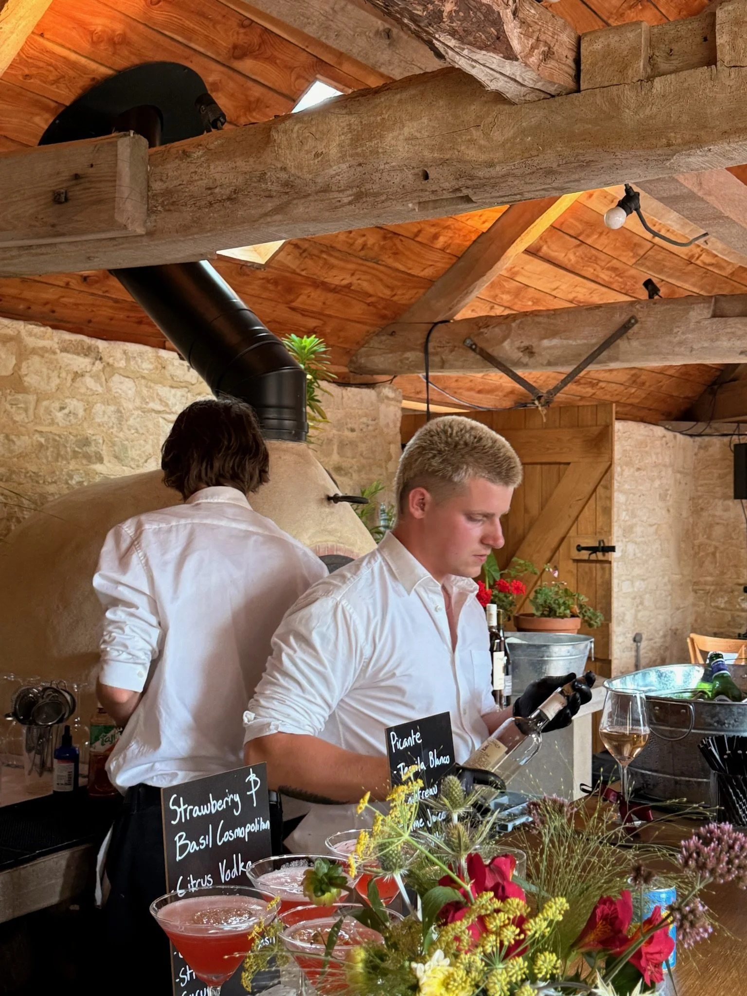 Bartenders preparing drinks behind a bar with floral decorations, a stone wall, and wooden beams in the background.