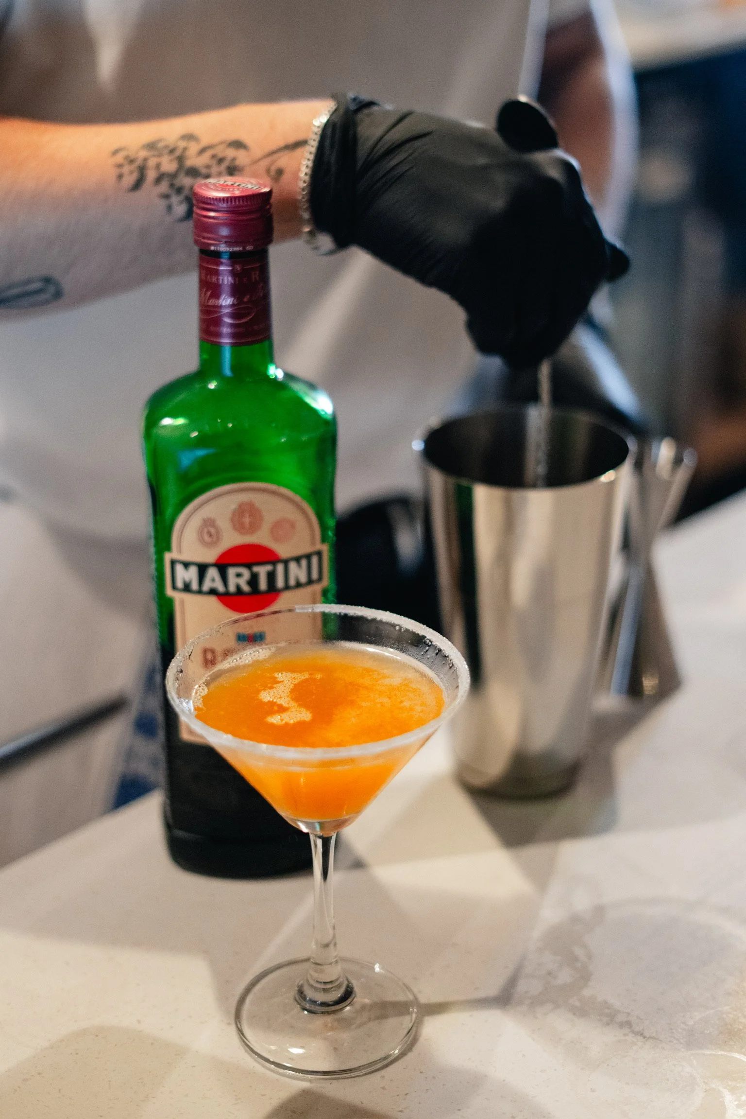 A bartender making a cocktail with a shaker, a bottle of Martini, and a martini glass with an orange drink