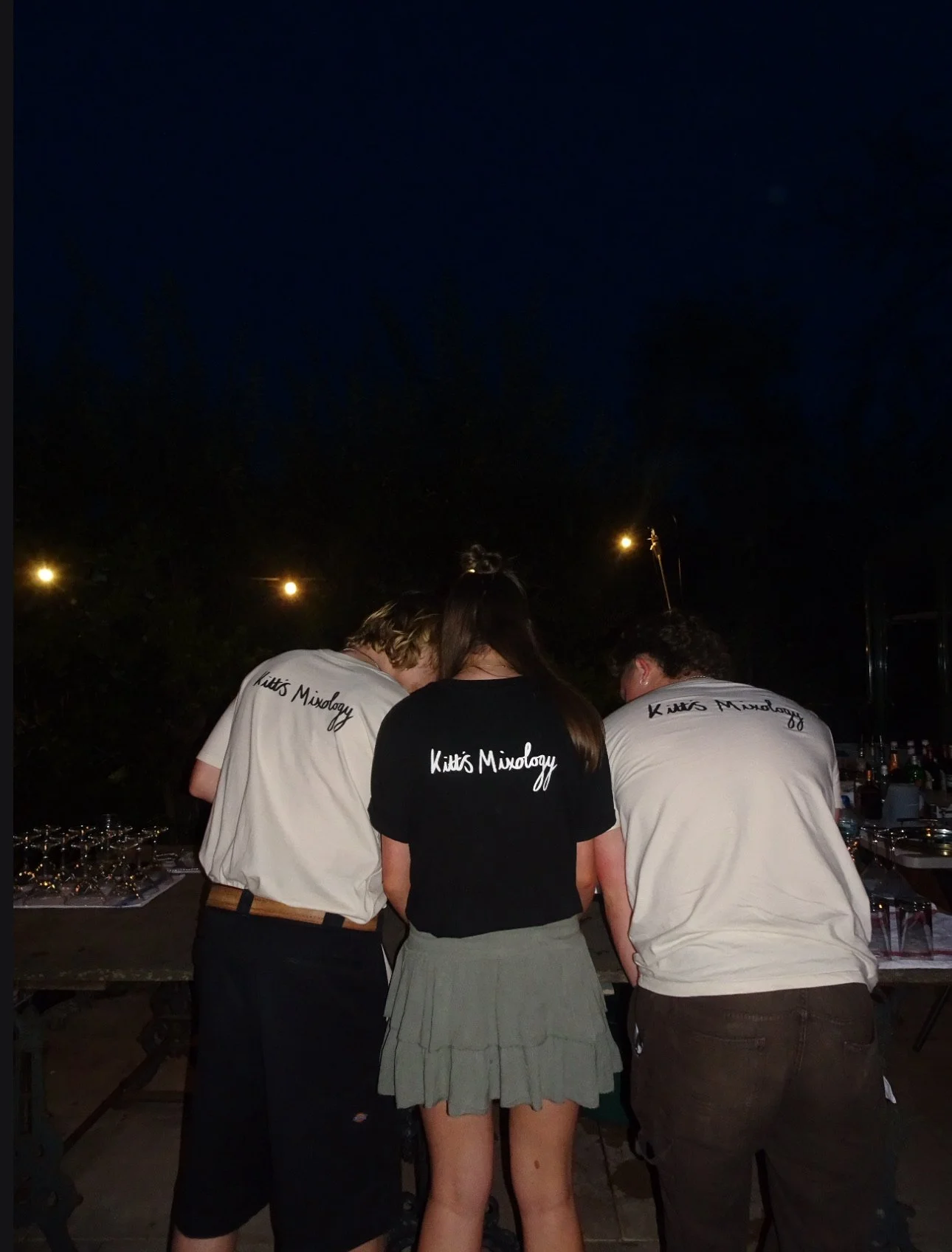 Three people, two men and one woman, look at a table with drinks and glasses outdoors at night. They are wearing shirts that say 'Kitt's Mindogy'.