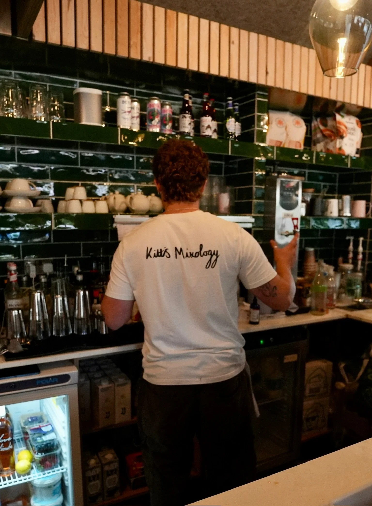 Barista working behind a counter in a restaurant or cafe, facing away, wearing a white T-shirt with 'Kitt's Mixology' written on the back, with shelves of glasses, bottles, cups, and bar equipment behind him.