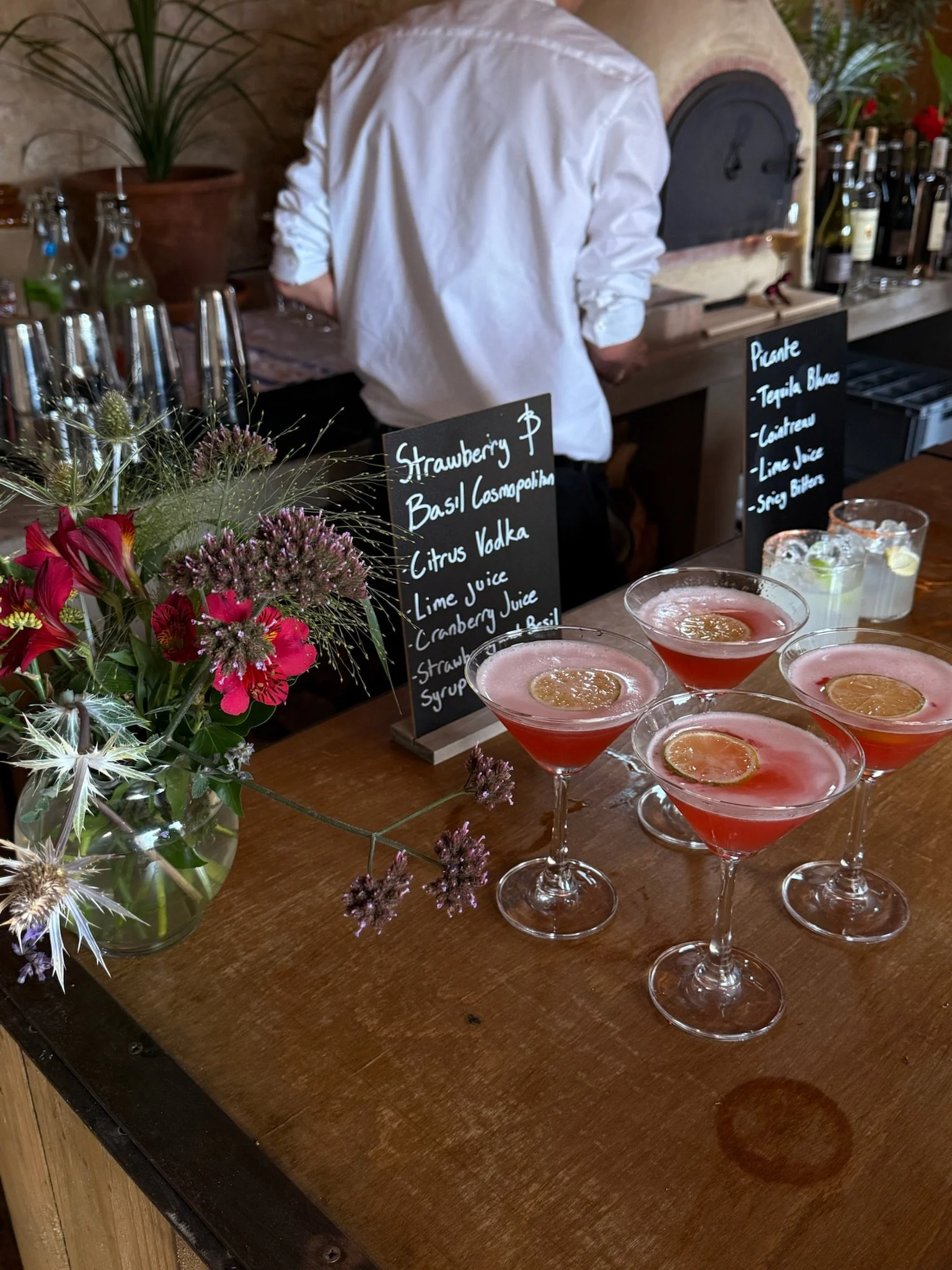 A bar counter with four pink cocktails garnished with lemon slices and a small floral arrangement. Two black chalkboard menus displayed behind the cocktails list ingredients for drinks.