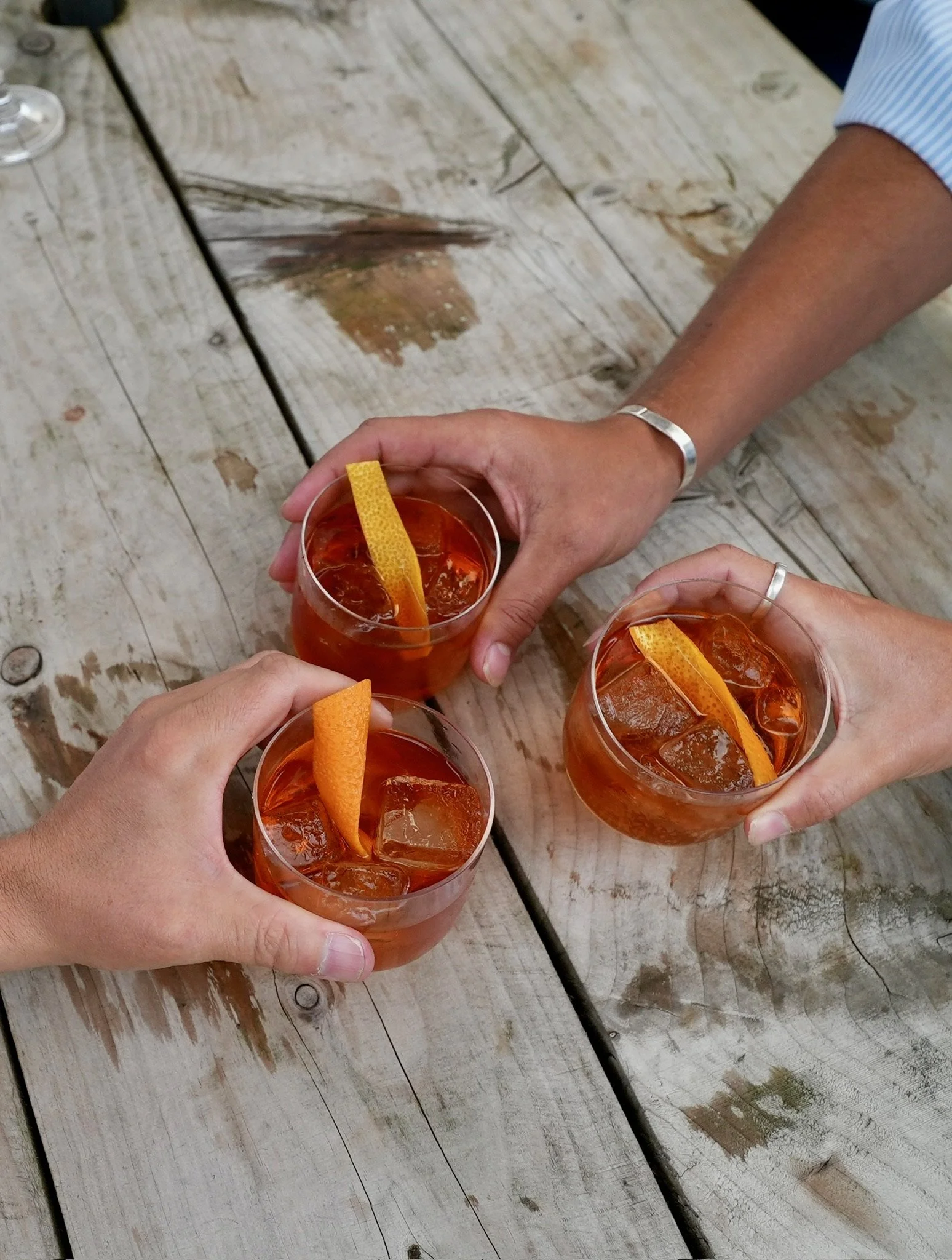 Three people holding glasses of an orange-colored drink with ice and orange peel garnish, seen from above on a rustic wooden table.