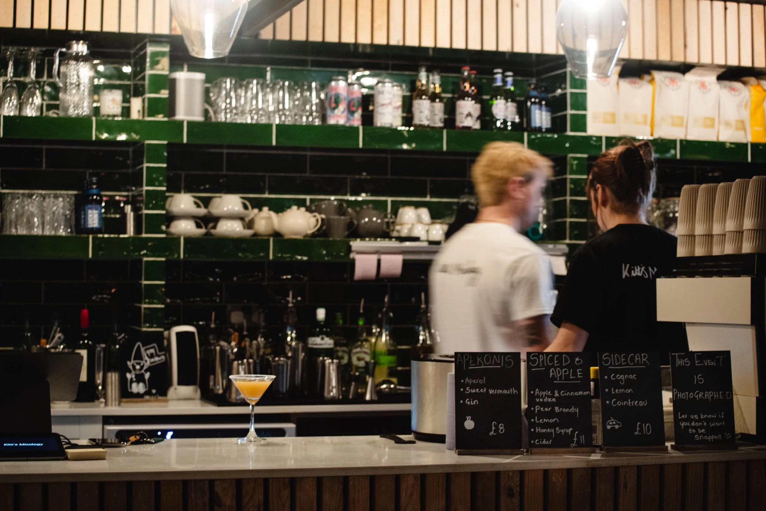 A bar or cafe counter with menus, glasses, and bottles behind it, two people standing and talking, and green tiled shelves with cups, glasses, and bottles.