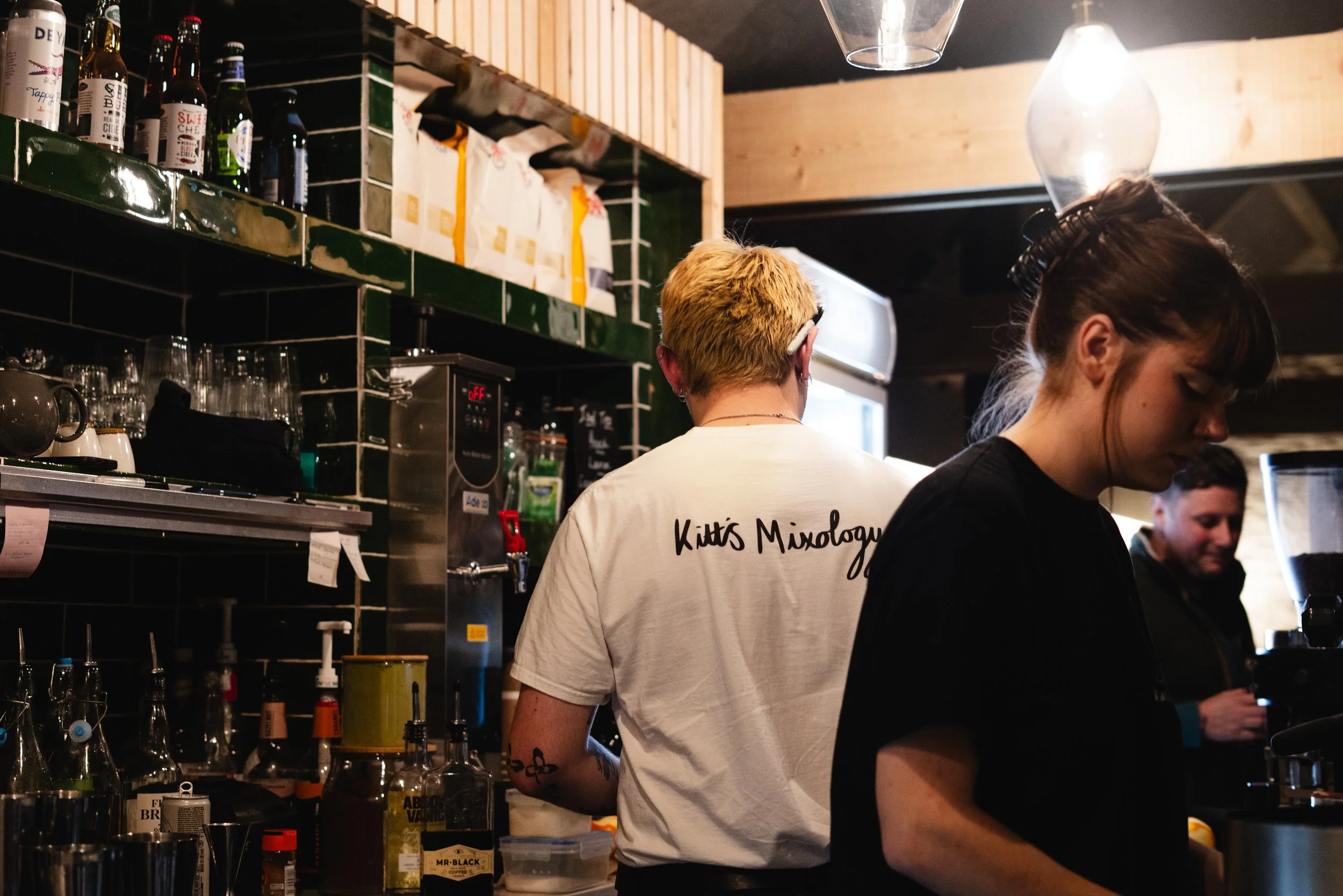 People working behind the bar at a cafe, with bottles, glasses, and bar equipment on the counter and shelves.