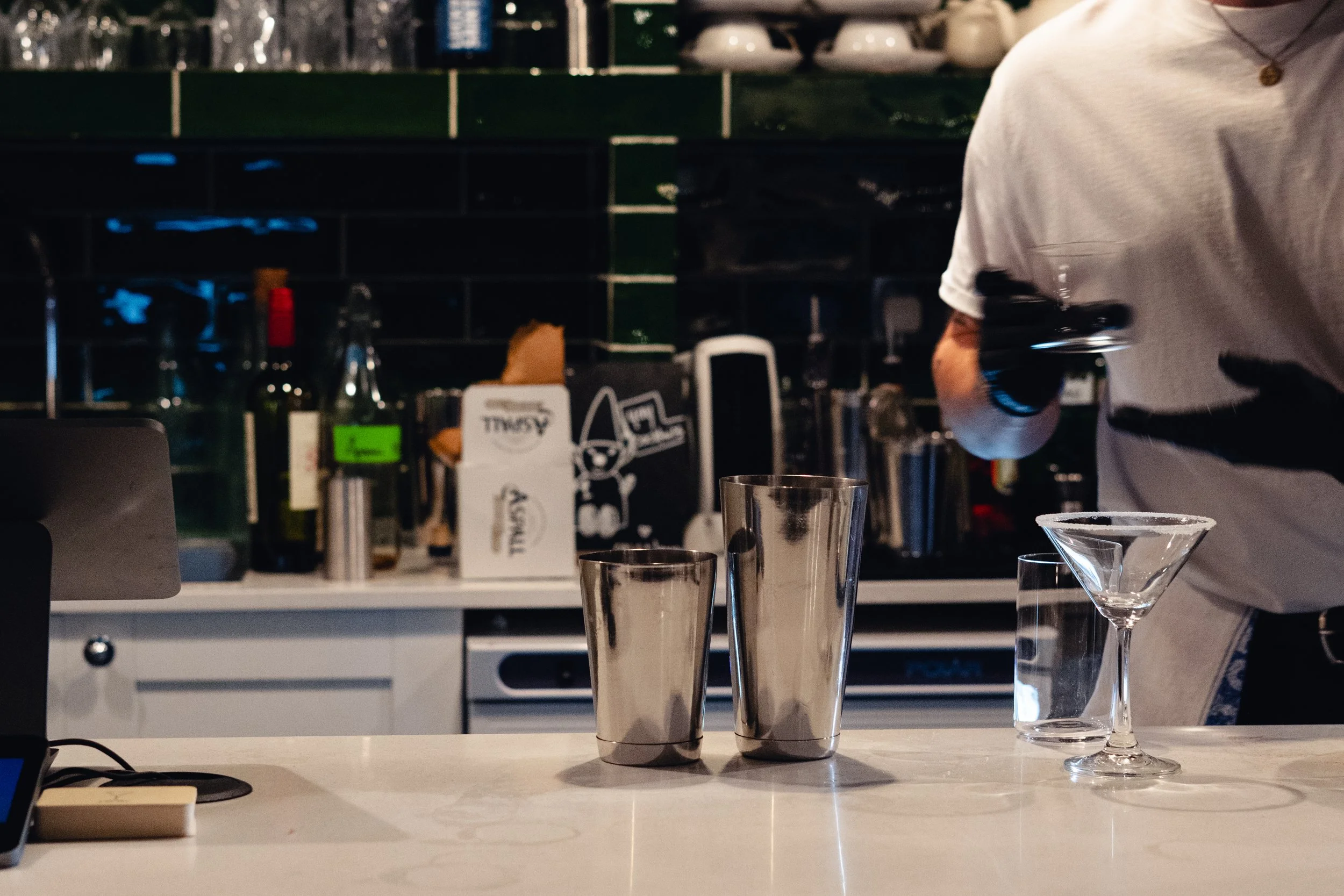 A person in a white shirt pouring a drink into a martini glass on a kitchen counter with two metal cocktail shaker and an empty glass nearby. The kitchen has a dark tiled backsplash and various bottles and objects on the counter in the background.
