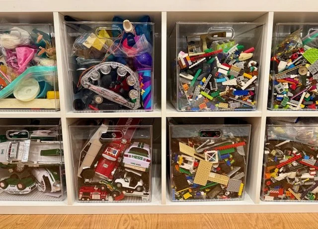 Decorative kitchen shelves filled with white milk bottles, small potted plants, glass jars with red decorative balls, miniature yellow cars, wooden utensils in containers, cookbooks, teacups with 'B,' 'A,' 'C,' and 'I' on them, and a set of nested mixing bowls.