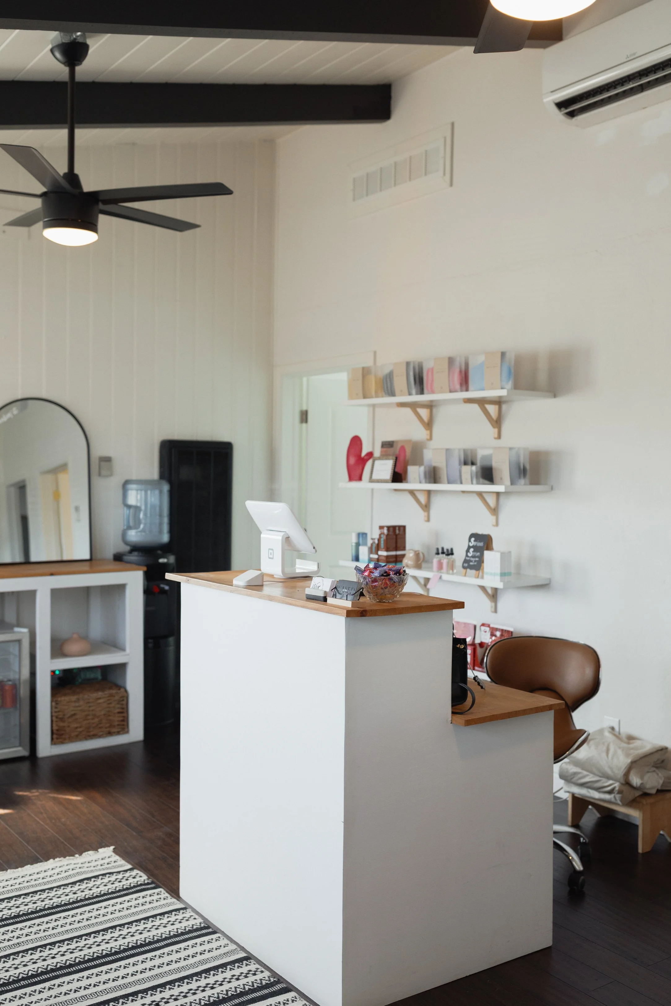 Interior of a cozy shop or reception area with a white counter, brown leather chair, wall-mounted shelves with products, a water cooler, and ceiling fans.