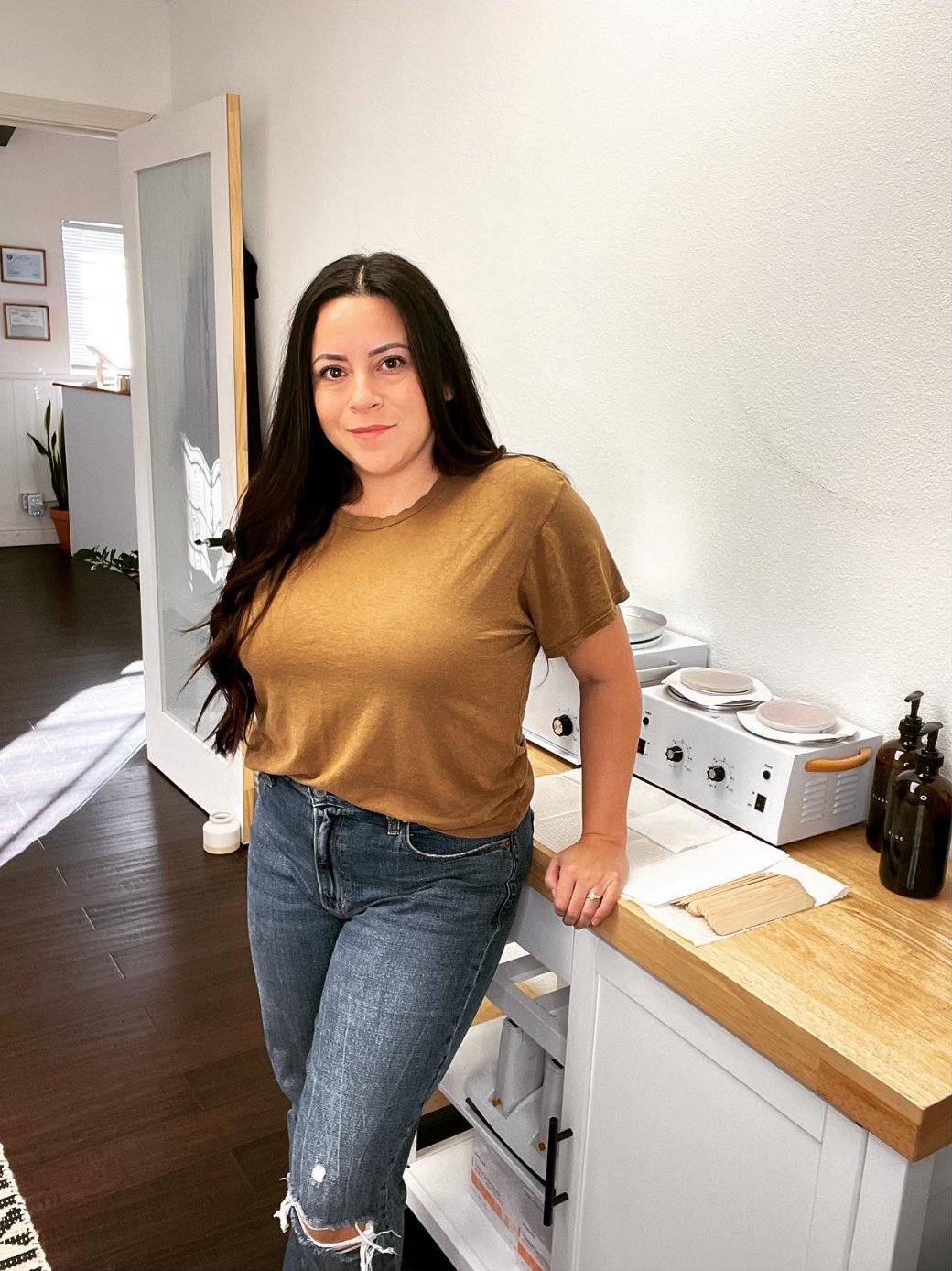 A woman with long dark hair, wearing a gold t-shirt and ripped jeans, standing in a kitchen.