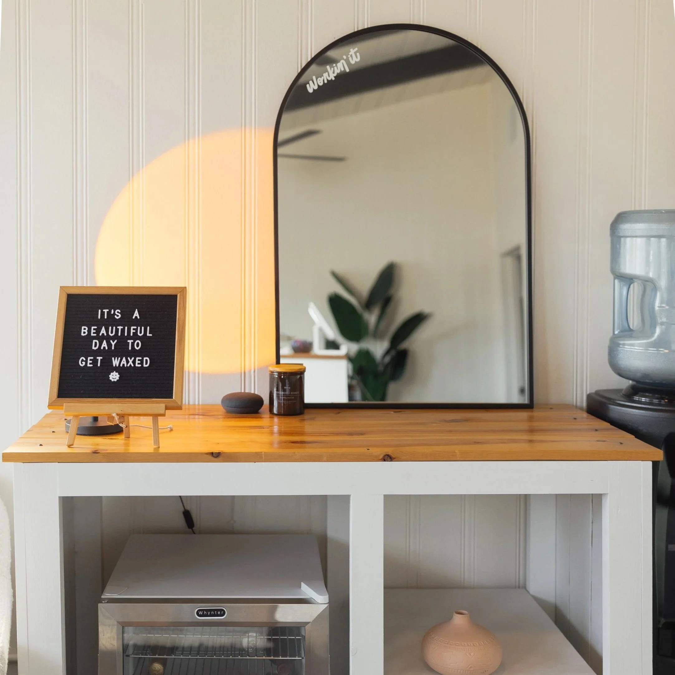 A wooden vanity table with a mirror, small decorative items, and a sign that reads 'It's a beautiful day to get waxed.' On the left, an orange light casts a glow. Below, there's a small appliance and a beige vase.