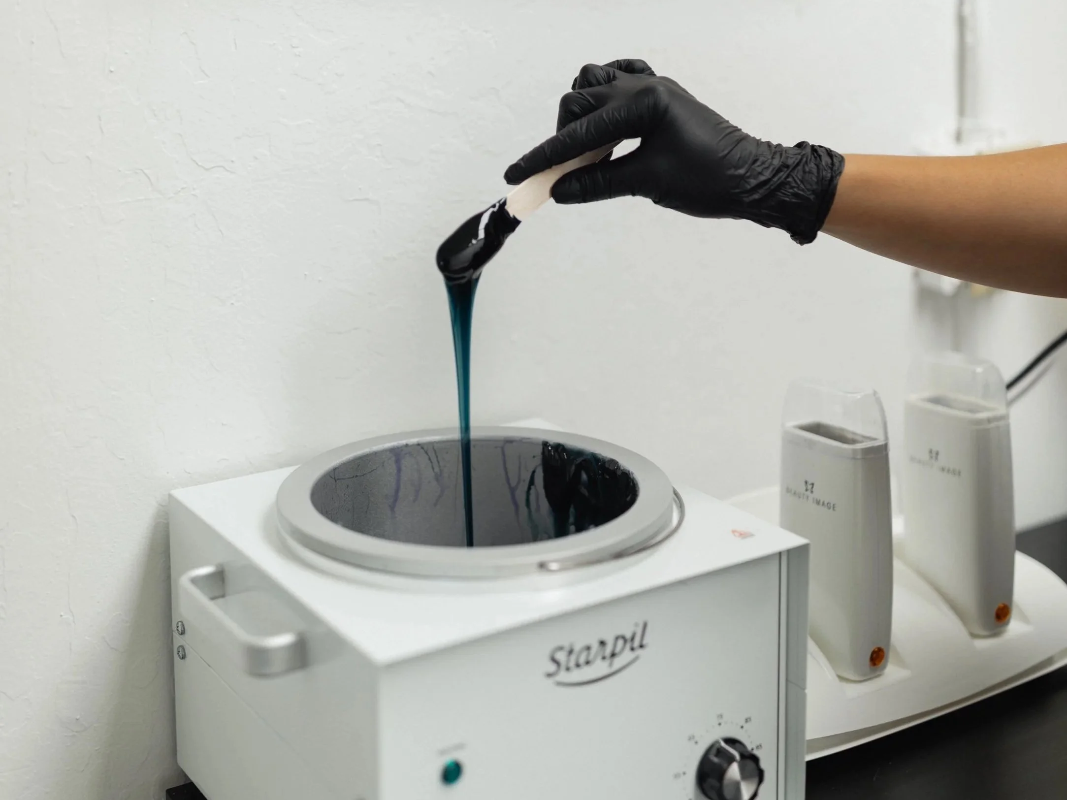 A person wearing black gloves is handling a sample with a black and white spatula over a laboratory heating device labeled 'Starpil', with two bottles of liquid or cream in the background.