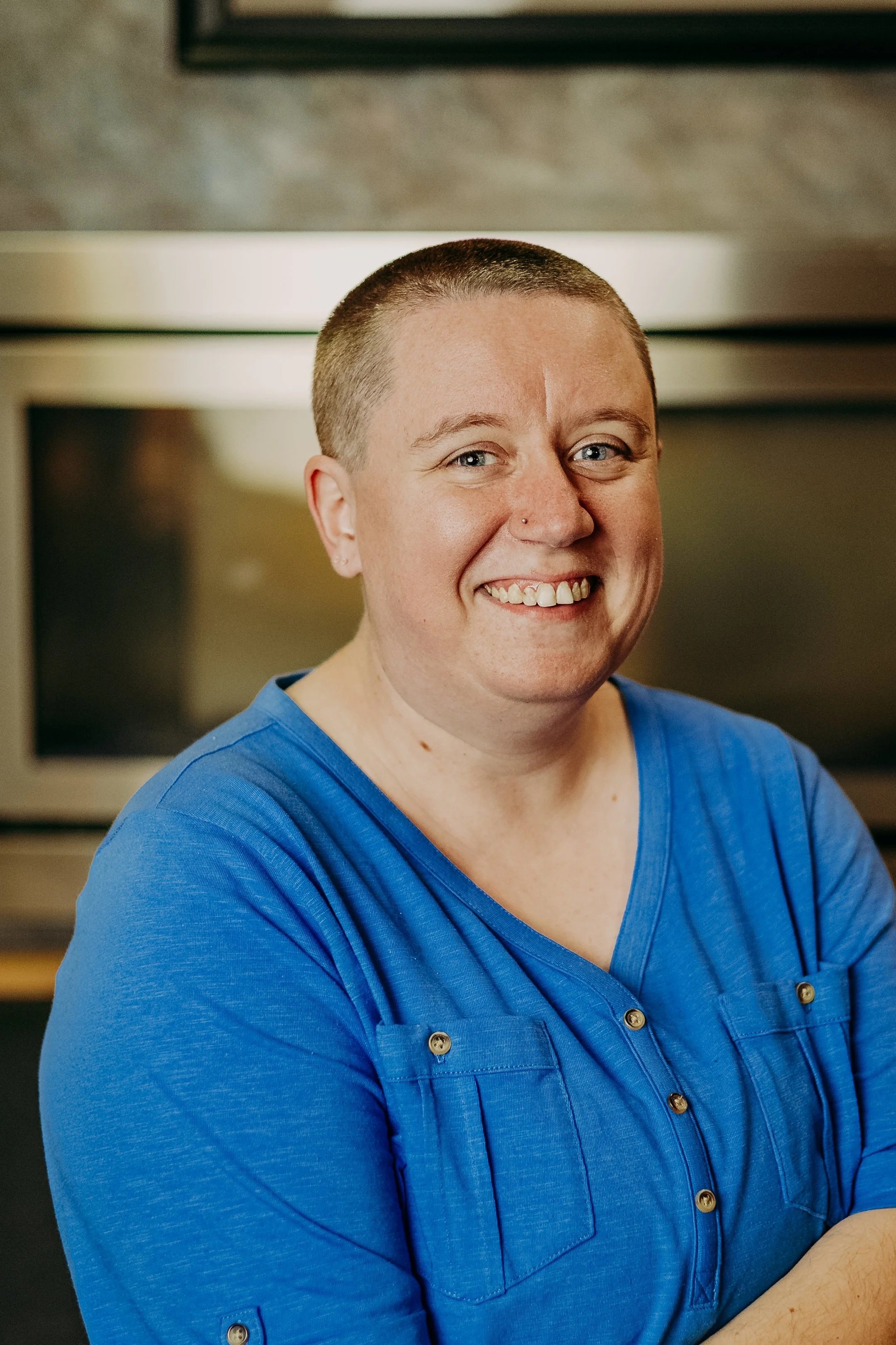 Close-up of a smiling person with short hair, wearing a blue shirt, in a kitchen setting.