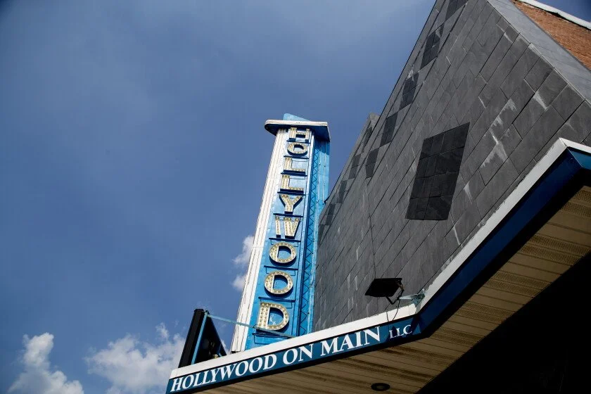 Close-up of a vintage Hollywood theater sign reading 'Hollywood' with blue neon lights, attached to a building with a black and gray exterior, against a background of blue sky with some clouds.