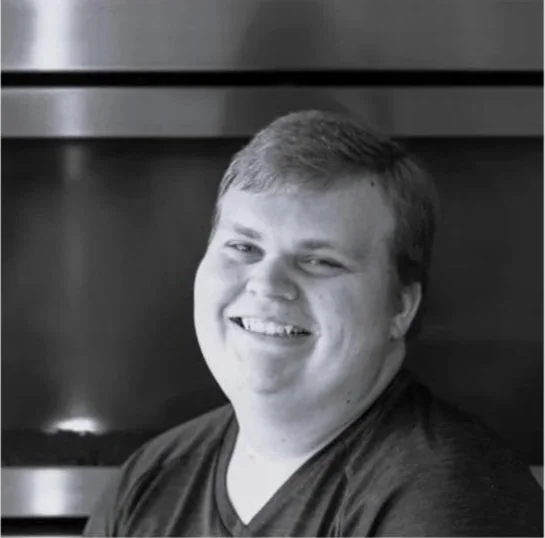 A young man with a smile, short hair, and wearing a dark shirt, in a black and white photo, sitting in front of a dark background.