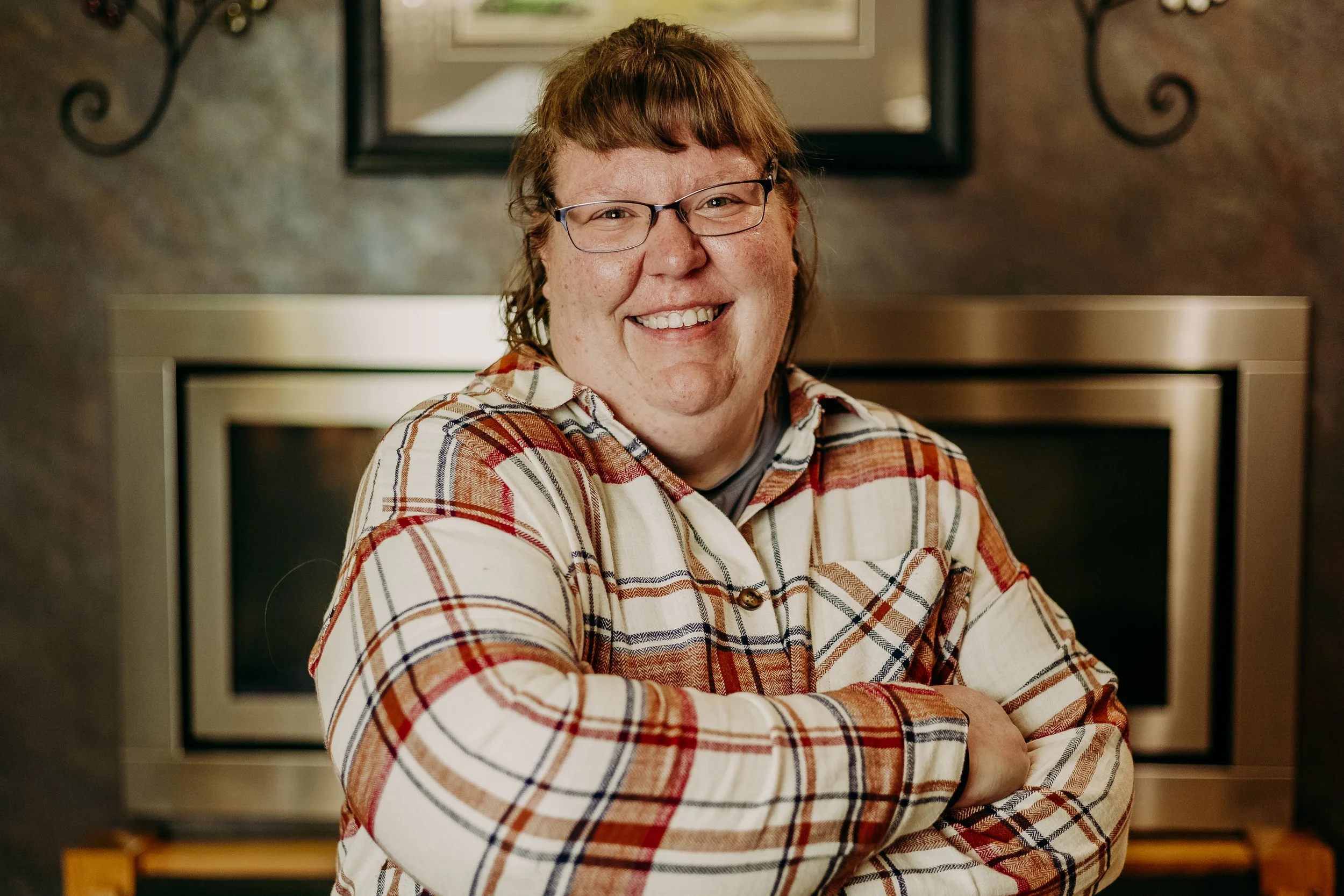 A person with short reddish-brown hair, glasses, and a big smile, wearing a plaid shirt, posing with arms crossed in front of a kitchen wall with a microwave oven.