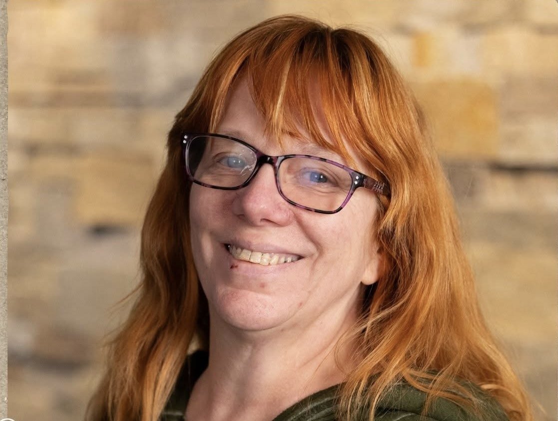 Portrait of a woman with long red hair, wearing glasses, smiling, standing in front of a blurred neutral background.