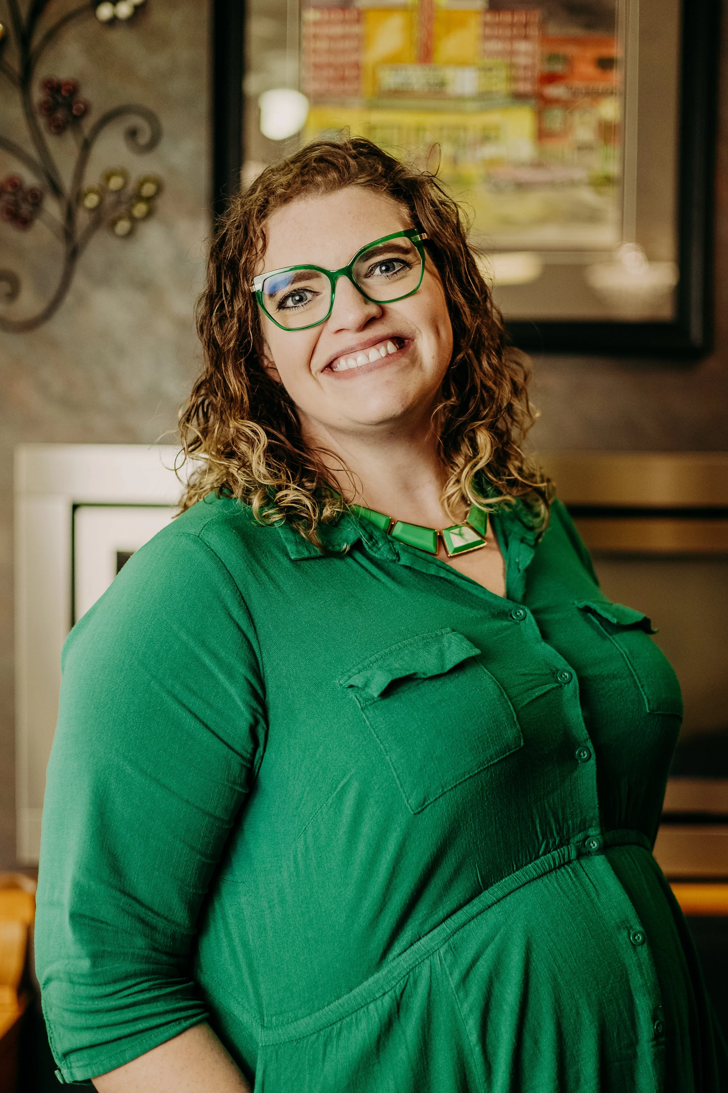 A woman with curly brown hair, wearing glasses with green frames and a green dress, smiling at the camera indoors with artwork and a textured wall in the background.
