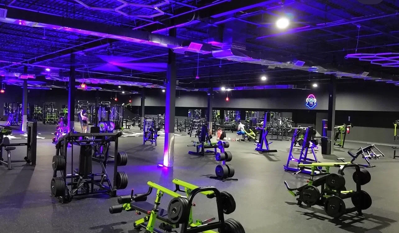 A modern gym with purple and black interior lighting, various strength training and cardio equipment, and a few people working out.