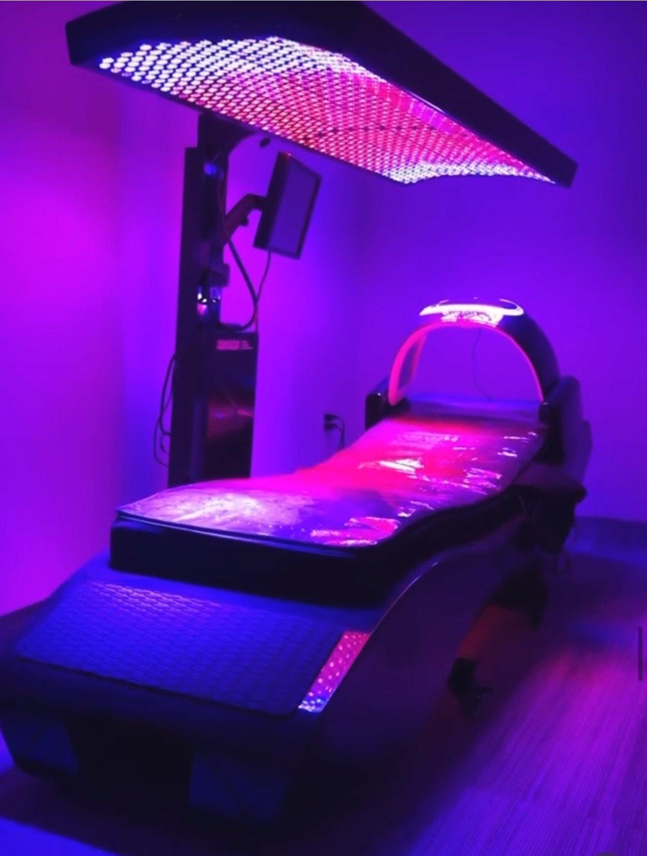 A medical or aesthetic treatment bed illuminated by purple and pink LED lights, with a control monitor attached to a stand beside it.
