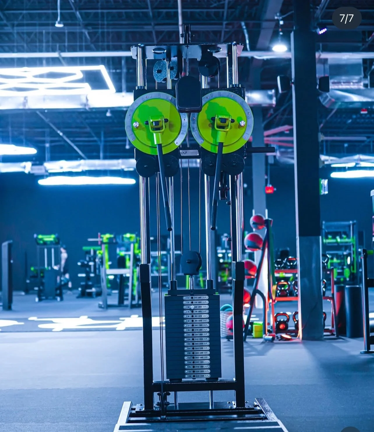 A cable machine with two green circular weight plates in a gym with various workout equipment and modern lighting.